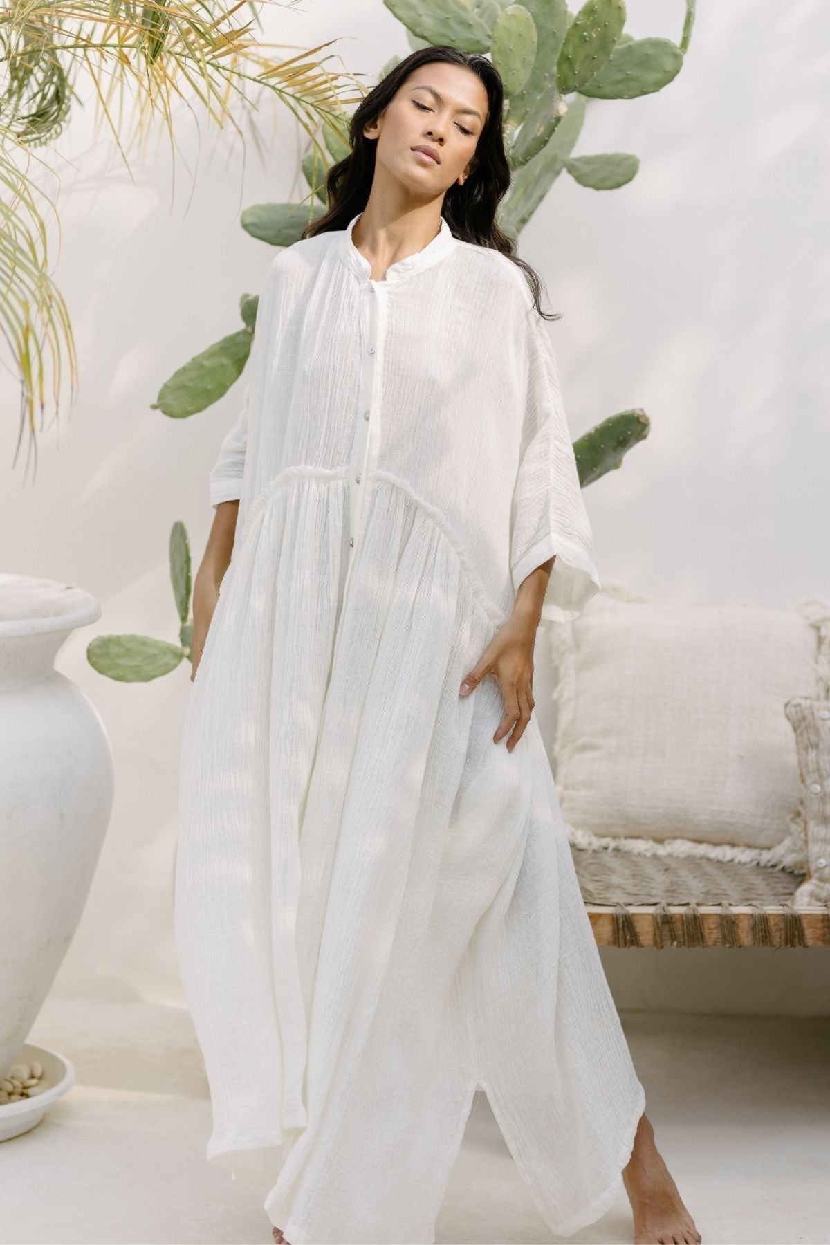 A person with long dark hair wearing a flowing Bona Dress (Cotton/Linen Crinkle) by Myrah Penaloza stands in a relaxed pose. They are in an outdoor setting with a whitewashed wall, large potted plants, and a woven bench with cushions. The ambiance is airy and serene, perfect for holiday season events.