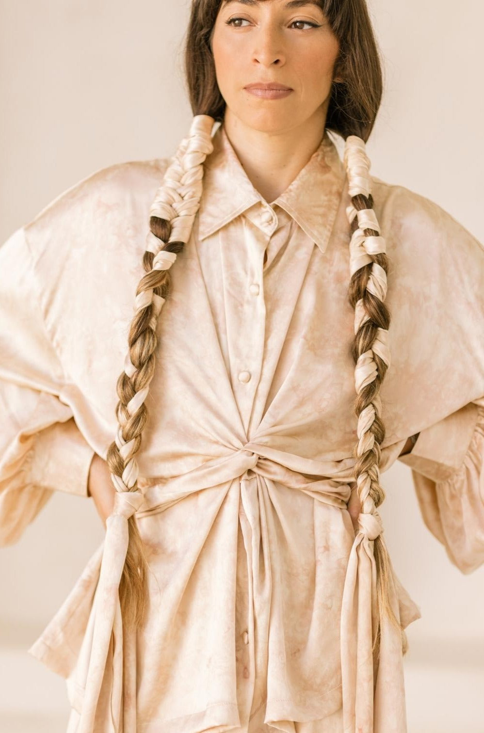 A woman with long braided hair stands confidently with her hands on her hips, dressed in the Inanna Top Only by Myrah Penaloza, a light-colored blouse made from 100% botanically dyed silk. Her braids are elegantly tied with fabric strips as she gazes to the side in a softly lit setting.