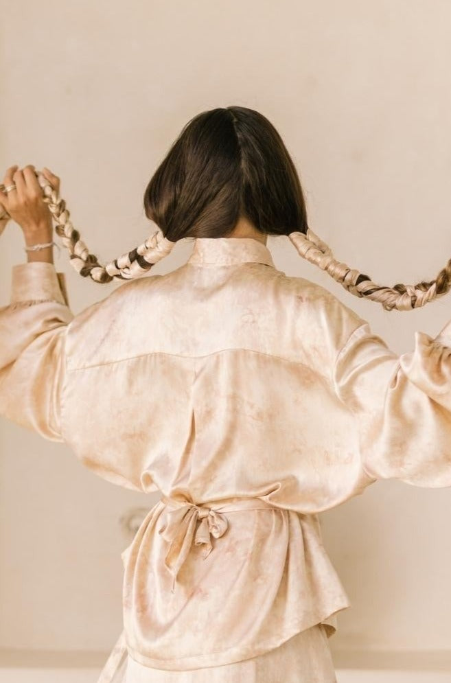 A woman with long, dark hair stands with her back to the camera, holding two braids in her hands. She is wearing the Inanna Top Only by Myrah Penaloza, made from 100% botanically dyed silk. The neutral beige background complements the scene.