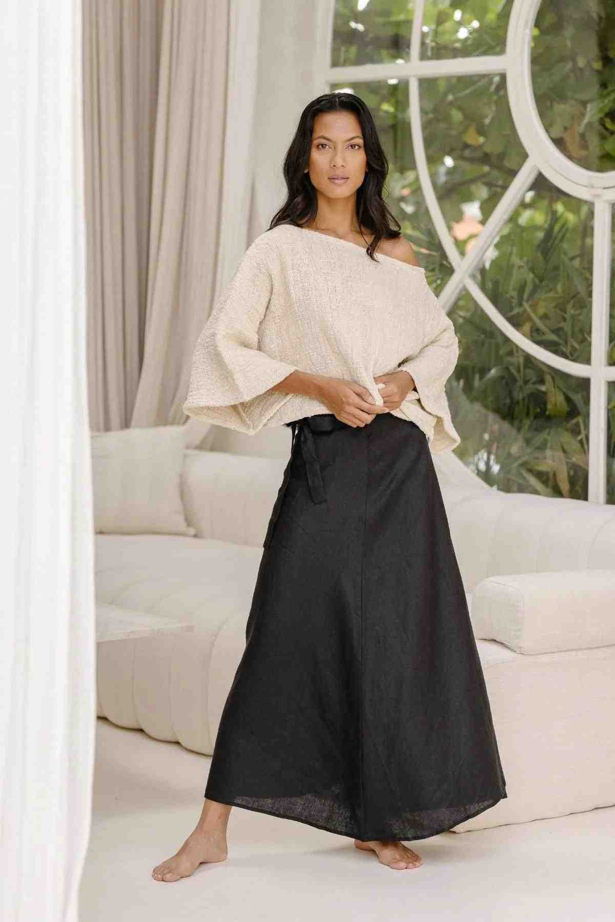 A woman stands barefoot in a brightly lit room with a large circular window, lush greenery visible outside. She is wearing the "Spirit Top & Shanti Wrap Skirt Set" by Myrah Penaloza, which includes a loose, off-white, 100% linen one-shoulder top with wide sleeves and a long, black adjustable wrap skirt. She has a calm expression and relaxed posture.