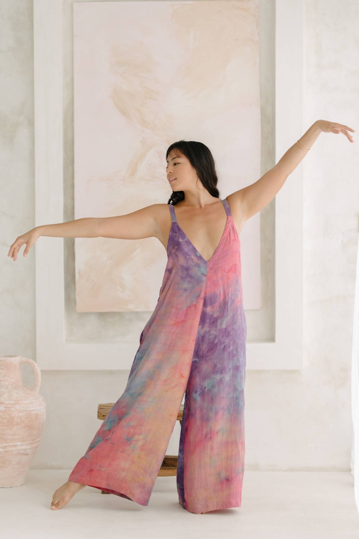 A person dressed in the vibrant Kundalini Linen Playsuit by Myrah Penaloza, featuring wide-leg bottoms and a tie-dye pattern, is striking a graceful, dance-like pose with arms extended. The background showcases a soft, pastel abstract painting and a large ceramic vase on the floor.