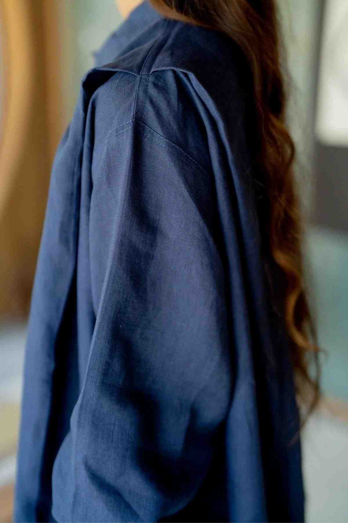 A person with long, wavy hair is wearing a loose-fitting, deep royal blue Tender Jacket made from 100% linen by Myrah Penaloza. The image captures them from the side, emphasizing the intricate details of the fabric and shoulder area of the jacket. Their face is not visible, adding an air of eclectic fashion to the scene.