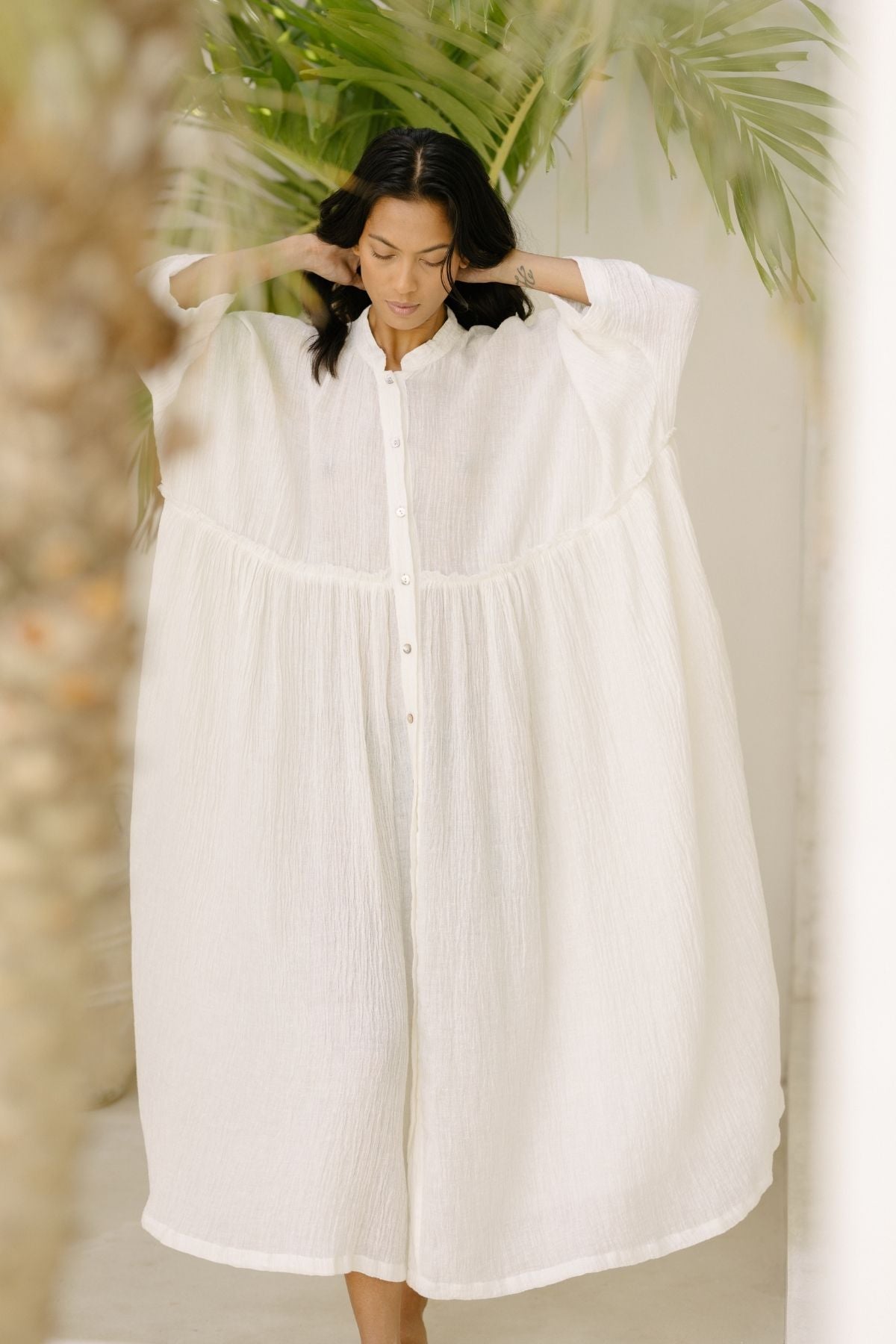 Indoors, a person stands gracefully in a loose, flowing Myrah Penaloza Bona Dress made from Cotton/Linen Crinkle. They adjust their hair with both hands as a lush green palm leaf in the background enhances the serene and tropical ambiance, perfect for holiday season events.