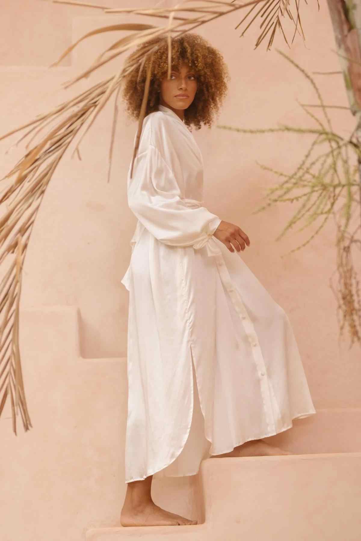 A person with curly hair is wearing a long, flowing Silk Inanna Wrap Gown by Myrah Penaloza, made of botanically hand-dyed silk, while standing barefoot on a set of light-colored stairs. They are looking towards the camera, surrounded by some dry plants and a smooth beige wall.