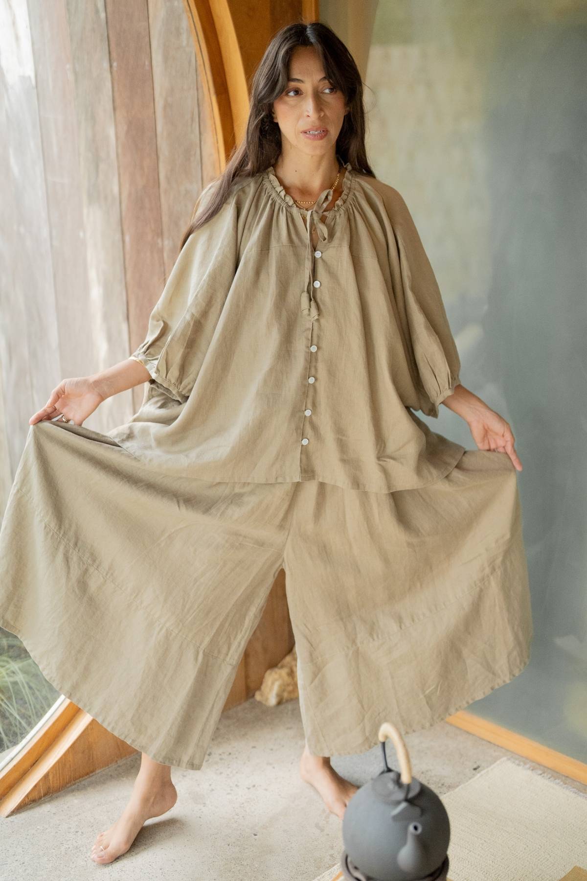 A woman with long dark hair is standing indoors, wearing a loose-fitting, beige Linen Playa Top by Myrah Penaloza that features wide sleeves. She is holding the sides of her top out with her hands. The background includes a curved wooden frame and a dark teapot with a handle, epitomizing a versatile and stylish look.