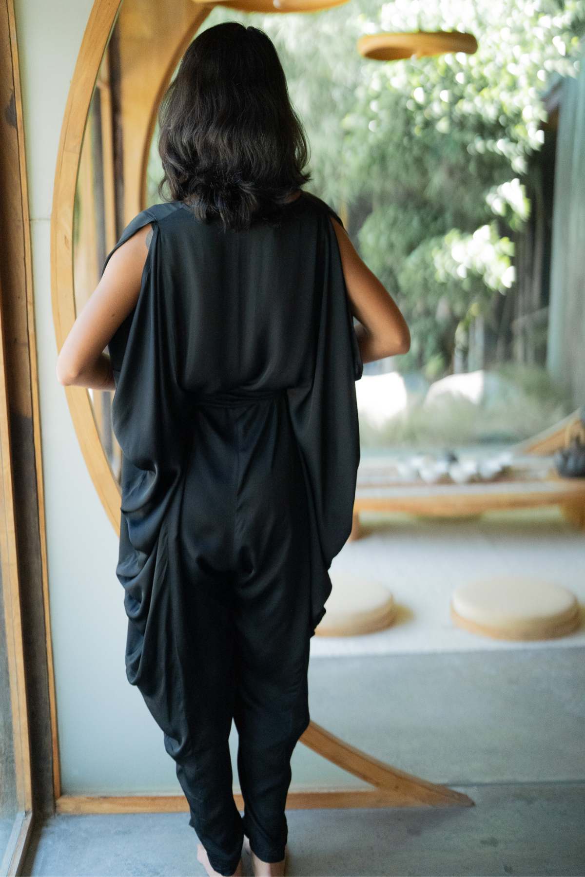 A person with long dark hair stands indoors, facing away from the camera, wearing a flowing Kuan Yin Playsuit by Myrah Penaloza. The 100% silk playsuit is botanically dyed in turmeric gold and made to order. They stand near a large window offering a view of greenery and softly lit round structures outside.