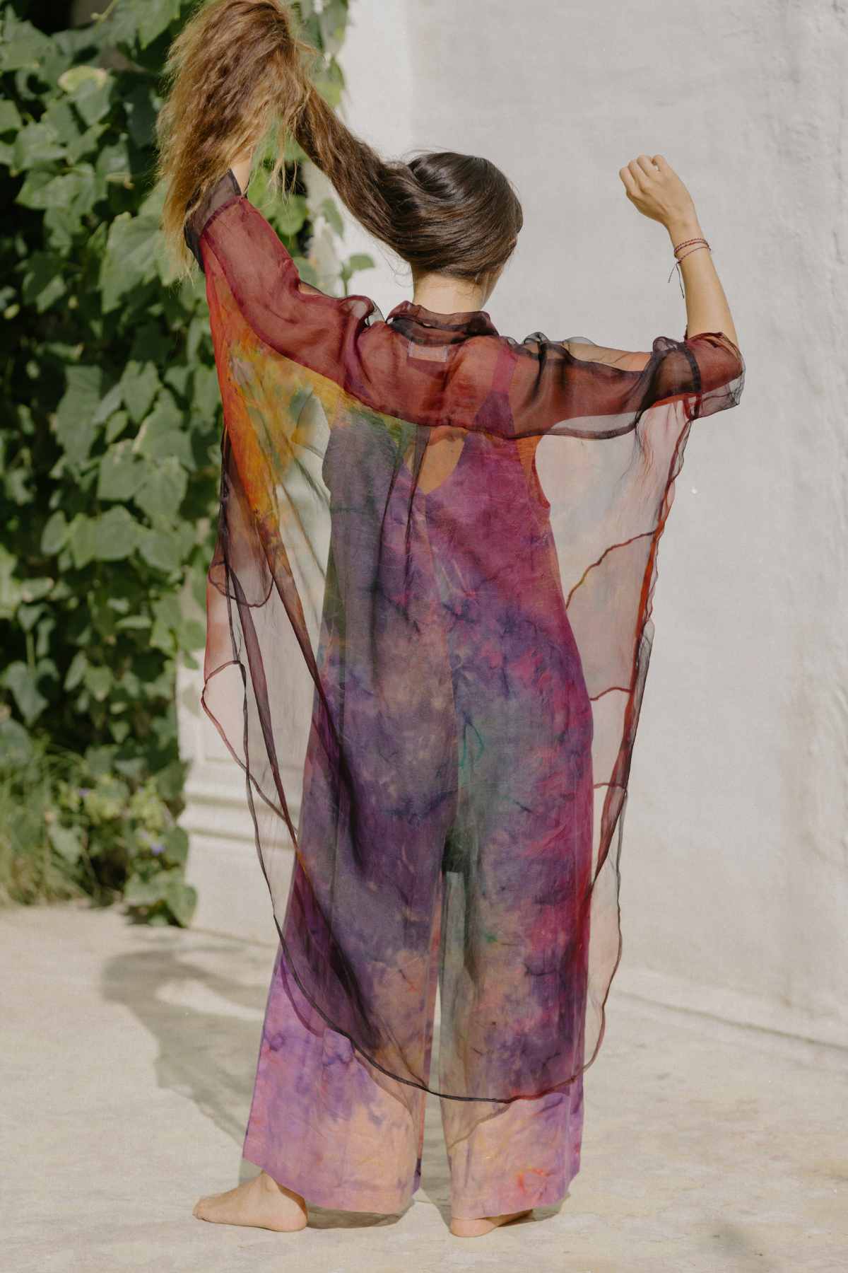 A person stands barefoot with their back to the camera, wearing an oversized fit Rainbeau Chiffon Silk Suka Top by Myrah Penaloza. The colorful translucent outfit is complemented by a flowing shawl as they extend their right arm upward, holding up their ponytail. On the left side, green foliage is visible against a light-colored wall.