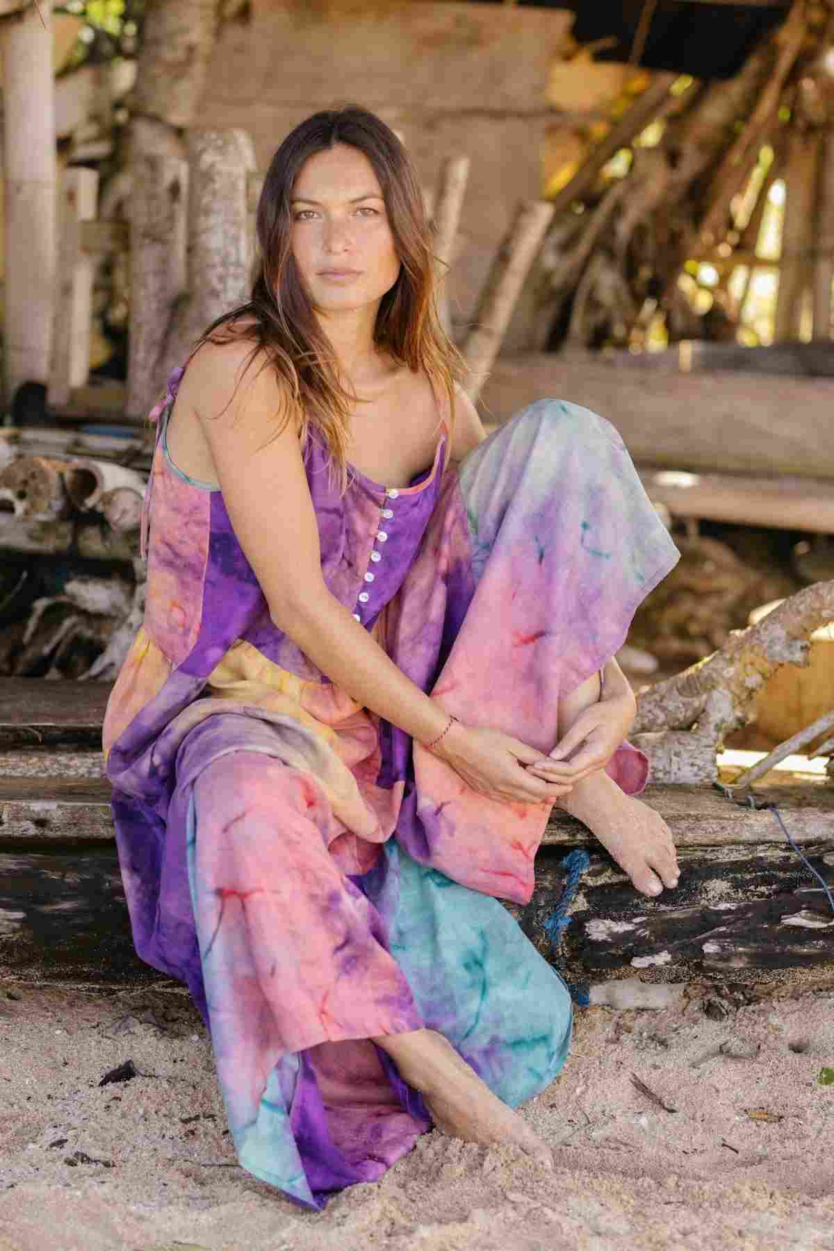 A woman sits on a wooden structure near the beach, wearing the vibrant La Mexicana Playsuit from Myrah Penaloza. She is barefoot and surrounded by driftwood and sand, her expression serene.