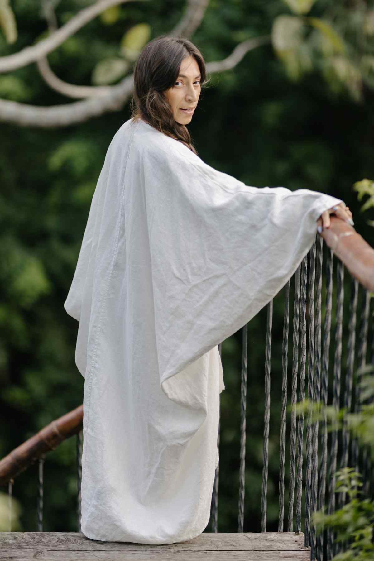 A person with long dark hair stands on a wooden bridge in a lush green forest. They are wearing a flowing white Dao Robe (100% Linen) by Myrah Penaloza, made of stone-washed linen. The person is turned slightly, looking over their shoulder toward the camera. The background is filled with vibrant green foliage.