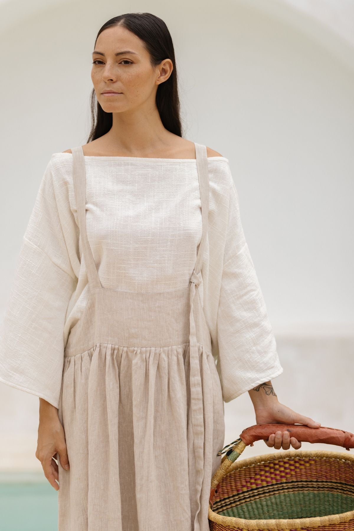 A woman with long dark hair, wearing Myrah Penaloza's light-colored May May Overall Skirt (Made To Order), stands holding a woven basket with a colorful base. She has a calm expression and is set against a neutral background with soft lighting, evoking the charm of a fairy garden.