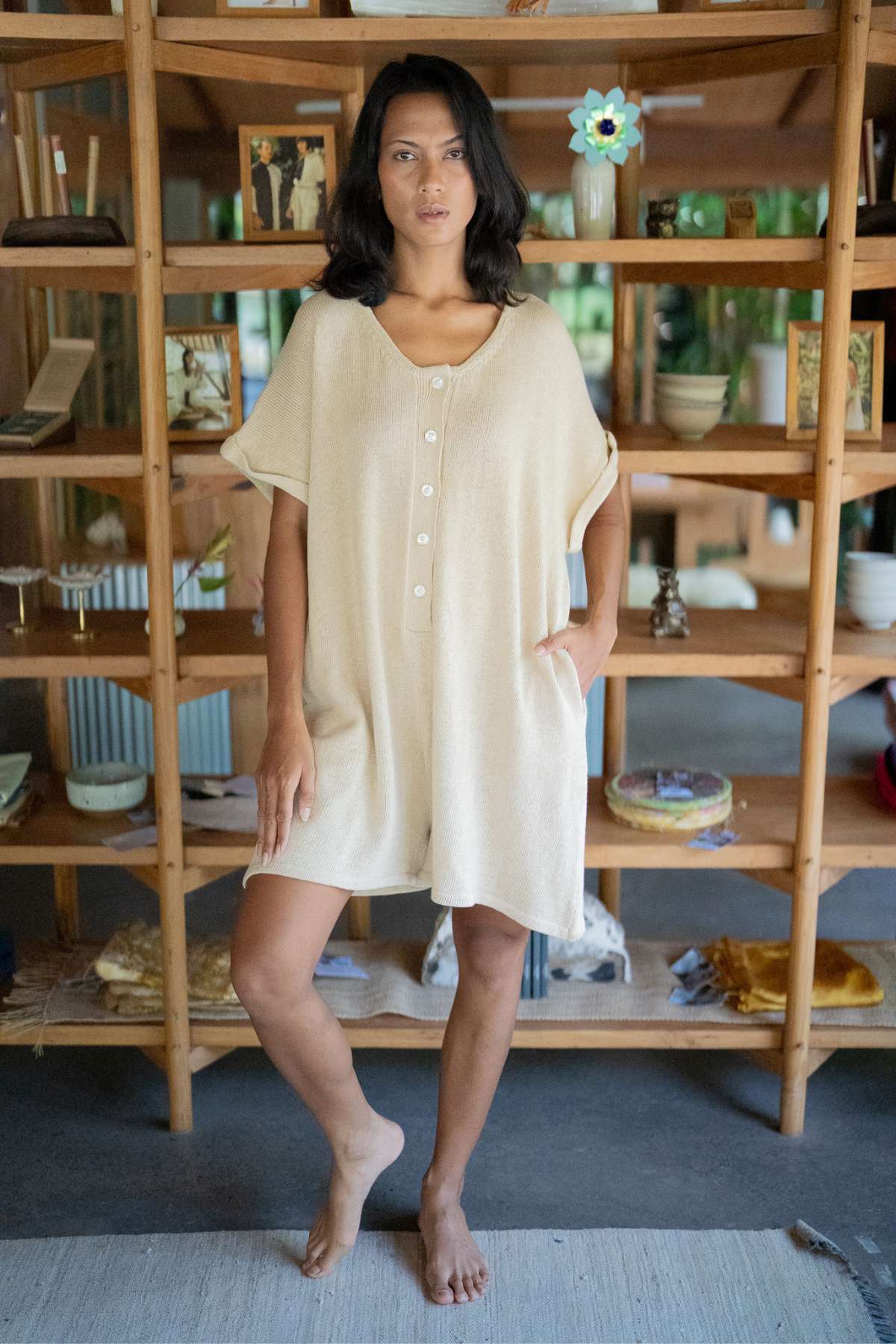 A person is indoors wearing the Henley Knitted Playsuit - BF by Myrah Penaloza, a light beige ensemble with a buttoned top and shorts. They are barefoot in front of a wooden shelf adorned with books, plants, and decorative items. This cozy cotton knit playsuit offers breastfeeding-friendly features.