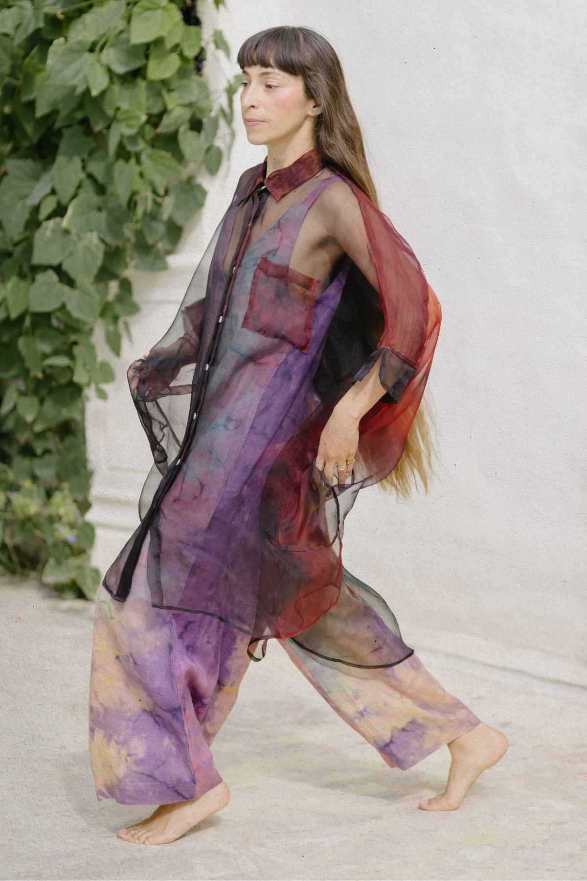 A person with long hair walks barefoot, dressed in an oversized fit Rainbeau Chiffon Silk Suka Top by Myrah Penaloza and matching wide-legged pants. The sheer, colorful outfit showcases a blend of red, purple, and green hues. They are outdoors, with a leafy plant in the background.