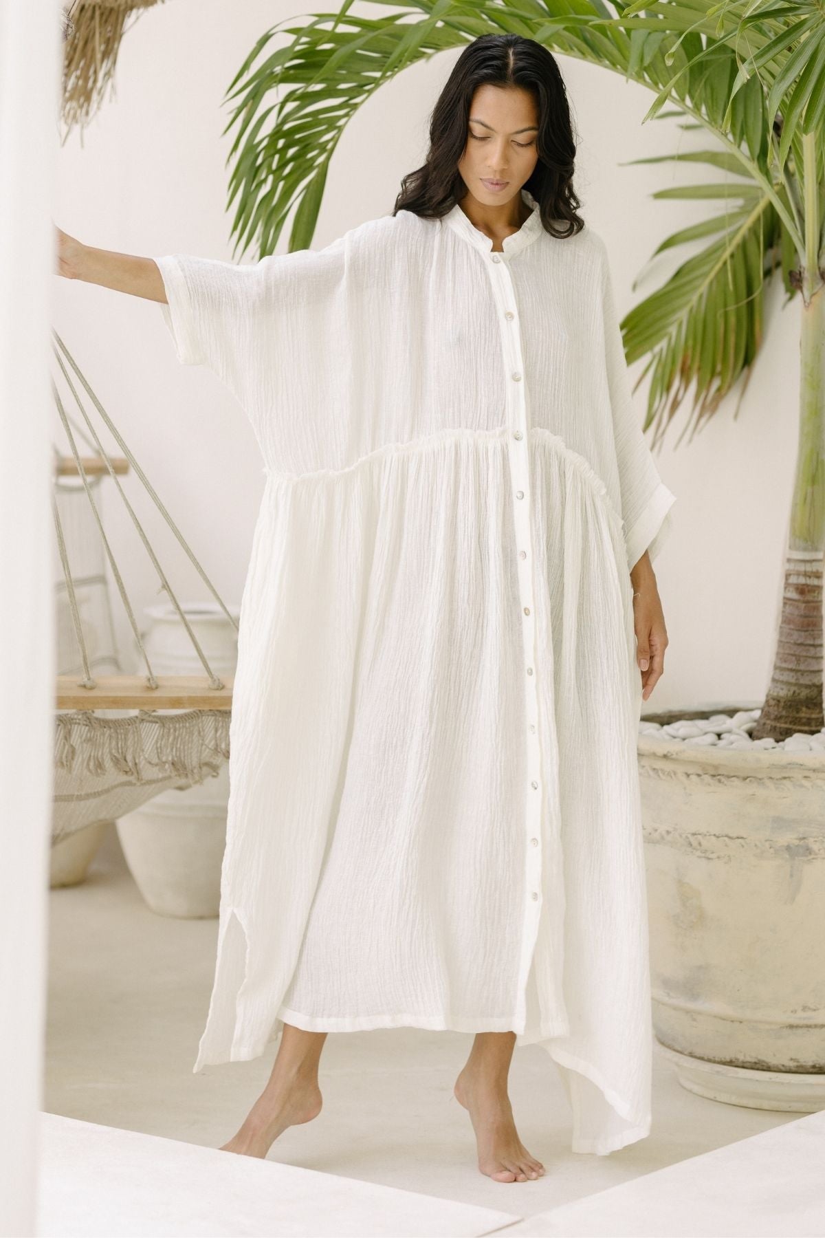 A woman dressed in a flowing, white Bona Dress (Cotton/Linen Crinkle) by Myrah Penaloza stands barefoot in an airy space with light-colored walls and large, green tropical plants. Her serene expression radiates peace as she lightly touches a hammock in the background, perfectly embodying the calm of holiday season events.