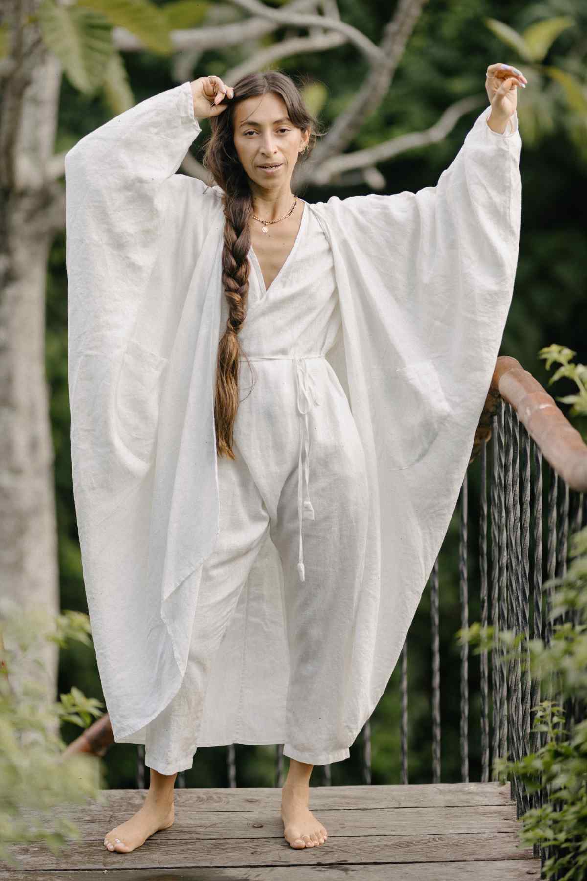 A barefoot person with long hair stands on a wooden platform, wearing a flowing Dao Robe made from 100% linen by Myrah Penaloza, featuring wide sleeves. They are outdoors, surrounded by greenery, with arms raised and one hand touching their head.