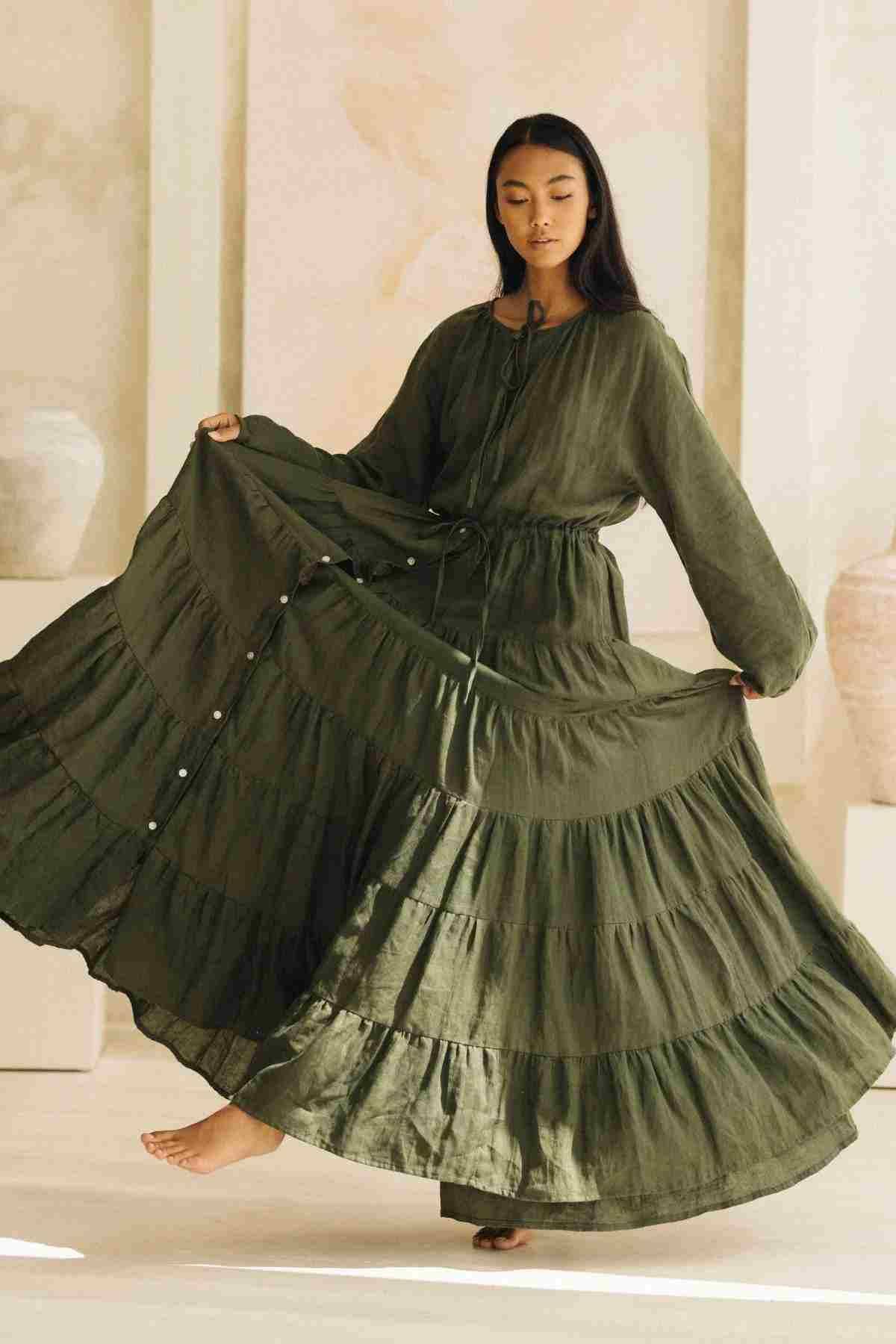 A woman gracefully poses indoors, barefoot and serene, wearing the La Mexicana Gown by Myrah Penaloza in a flowing green color. The gown features an adjustable waist, and she holds the sides of its voluminous skirt to showcase its fullness. Her long dark hair complements the soft, neutral tones of her surroundings, accented by large vases.