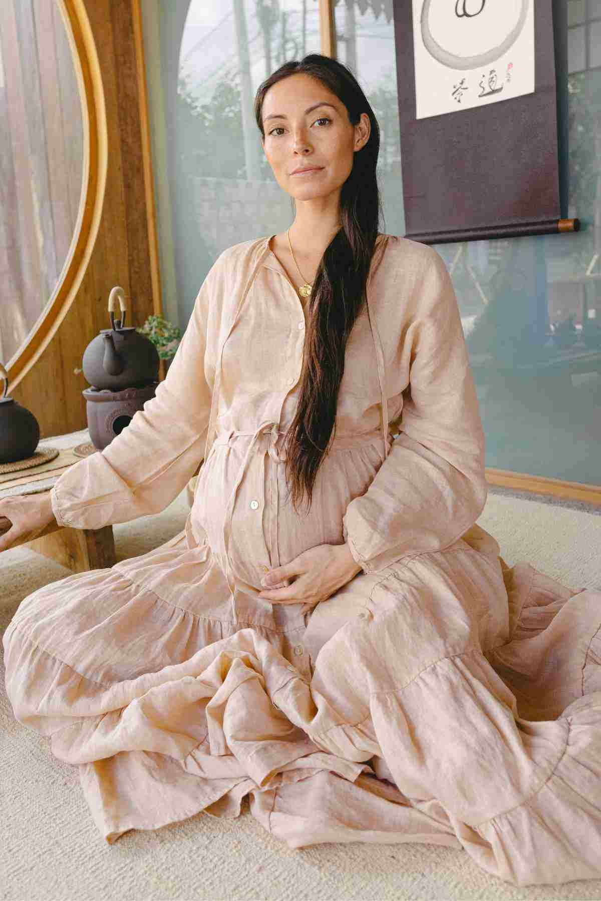 A pregnant woman wearing the La Mexicana Gown by Myrah Penaloza sits cross-legged indoors, gently resting her hands on her belly. She is surrounded by soothing, minimalist decor with natural light filtering through a round window.