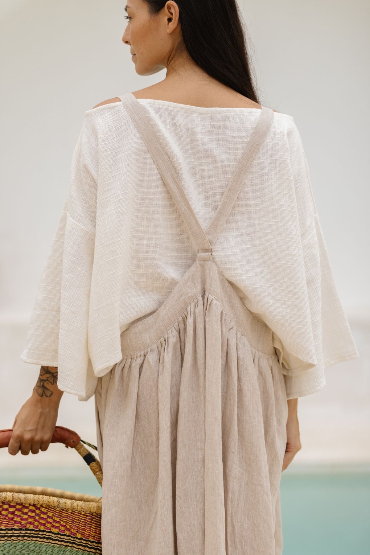 A woman with long, dark hair stands facing away from the camera, wearing a loose off-white crinkle linen top and the May May Overall Skirt by Myrah Penaloza. Her left arm rests on a colorful woven basket bag.