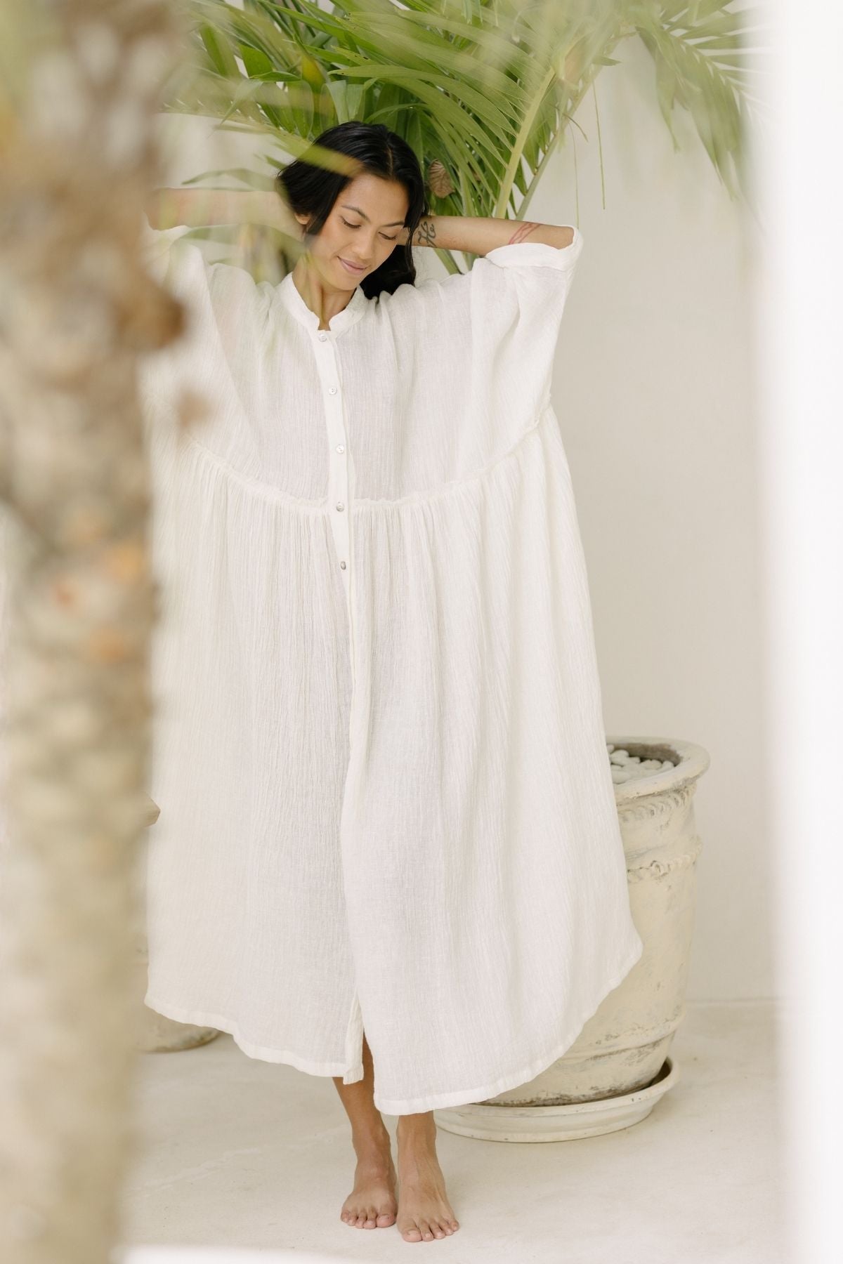 A person stands barefoot on a light-colored surface wearing a flowing, long white Bona Dress from Myrah Penaloza with button details. They have their arms raised, adjusting their hair. Green palm fronds are visible in the background. The Cotton/Linen Crinkle fabric enhances the serene and tropical vibe of the scene.