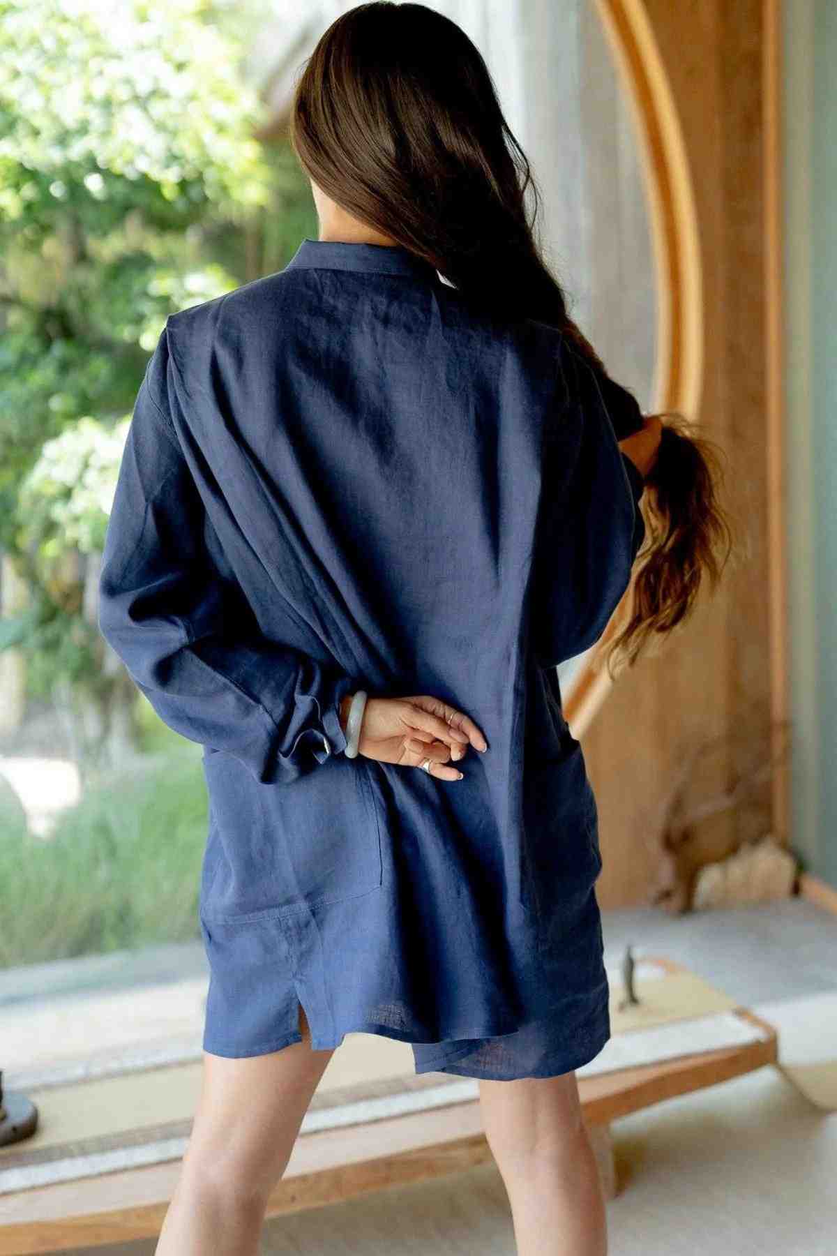 A person with long brown hair stands indoors with their back to the camera. They are wearing an oversized Tender Jacket (100% Linen) by Myrah Penaloza, holding their hair behind their back with one hand. A large, round window in the background offers a view of greenery outside, highlighting the eclectic fashion sense of their deep royal blue ensemble.