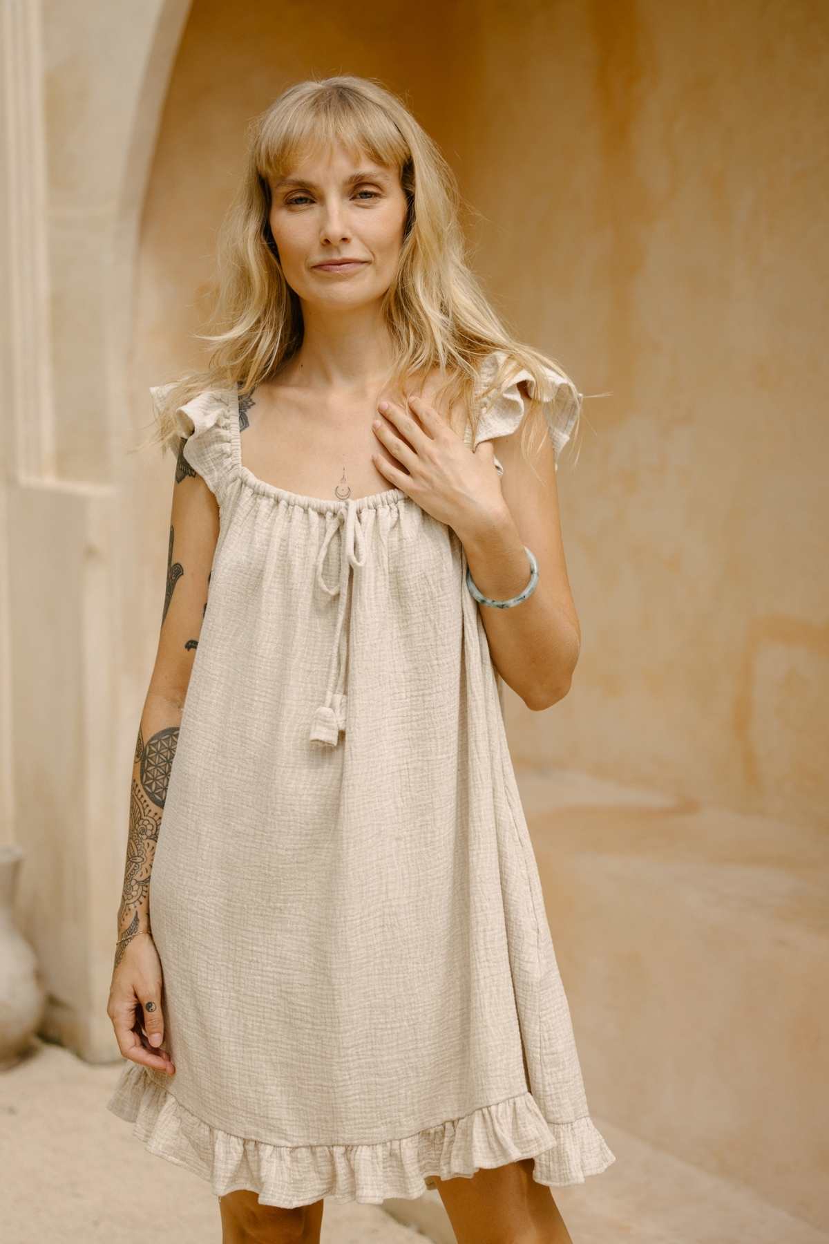 A woman with long blond hair and bangs stands in front of a beige wall, wearing the Myrah Penaloza Bumi Mini Slip Dress, which features ruffled straps and hem. She has tattoos on her arm and is gently touching her chest with her right hand while looking at the camera, perfectly embodying the essence of luxurious loungewear.