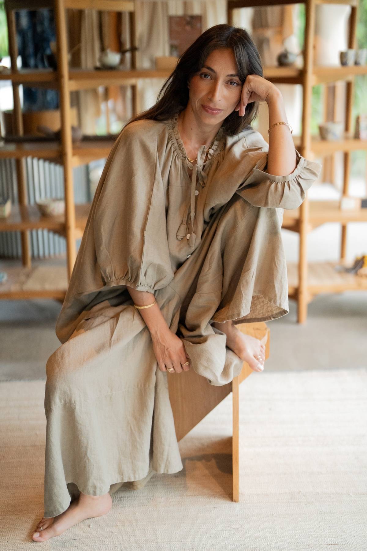 A woman is sitting on a wooden stool in a cozy, well-lit room with shelves filled with various items. She is wearing the versatile and stylish Linen Playa Top from Myrah Penaloza—a loose, beige piece made from 100% natural linen, complete with long sleeves. Her dark hair is styled naturally as she rests her chin on her hand, gazing thoughtfully at the camera.