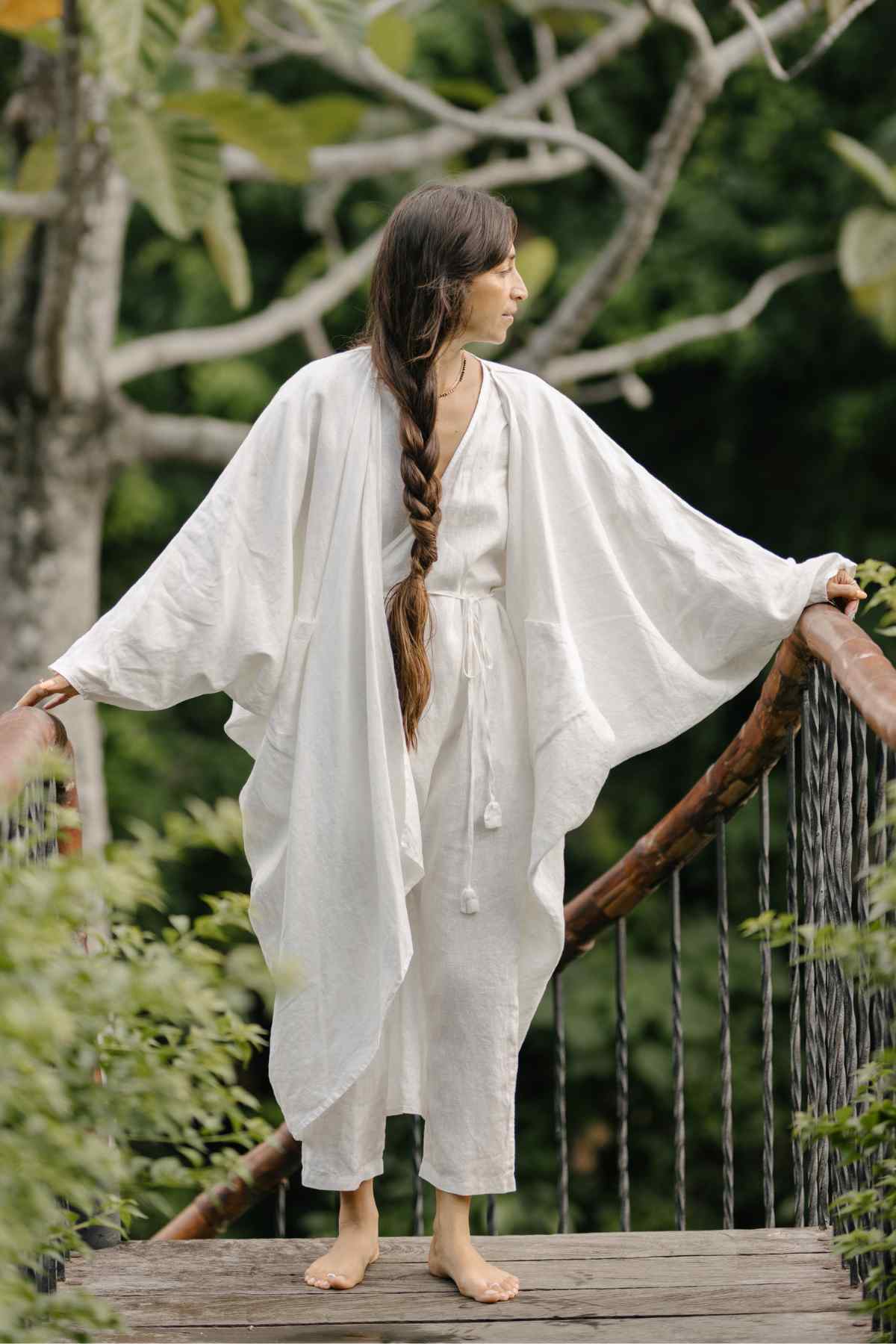 A barefoot person with long braided hair stands on a wooden bridge, draped in a flowing Dao Robe made of 100% stone-washed linen by Myrah Penaloza. They look to the side, surrounded by lush greenery, giving the scene a serene and natural vibe.