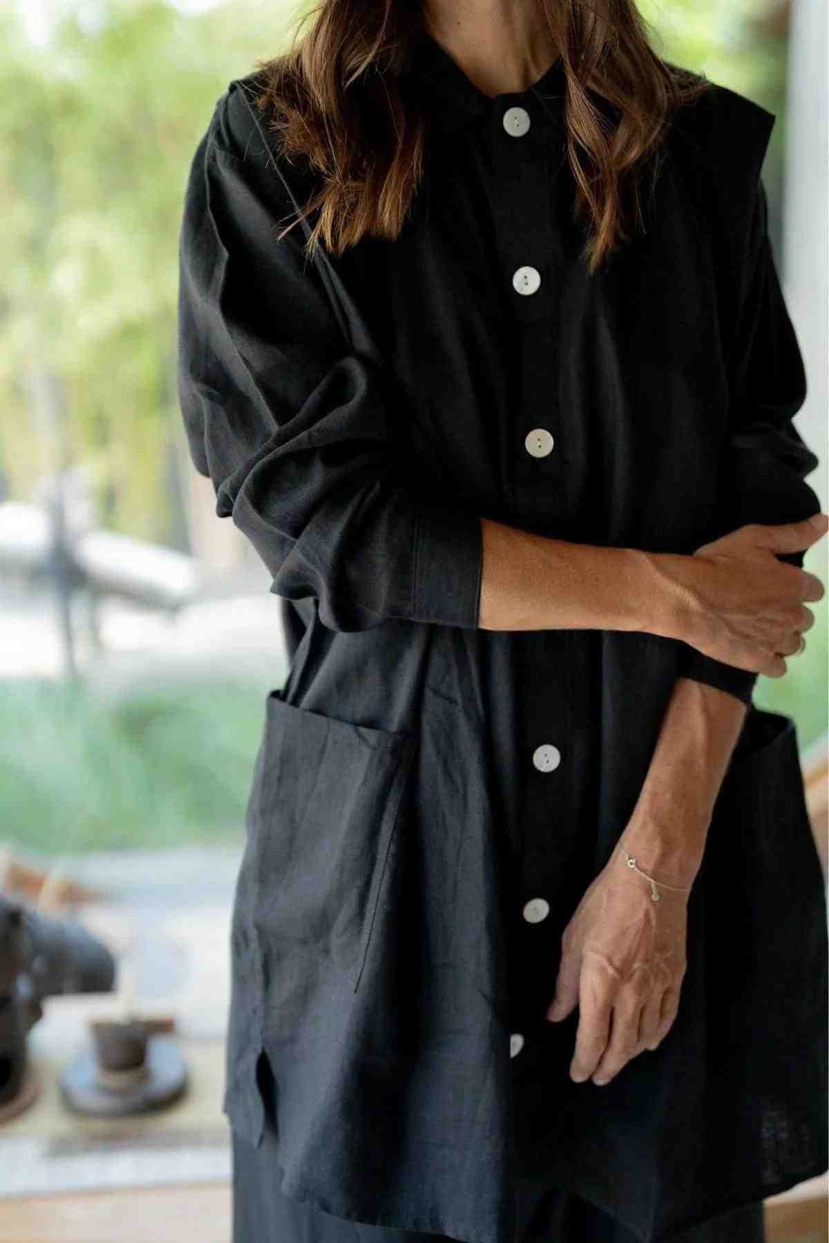 A person stands with one arm crossed over their body, wearing the Tender Jacket (100% Linen) by Myrah Penaloza, a loose-fitting black piece featuring white buttons and large front pockets, perfectly complementing their eclectic fashion sense. The background appears to be a blurry indoor setting with a hint of greenery visible through a window.