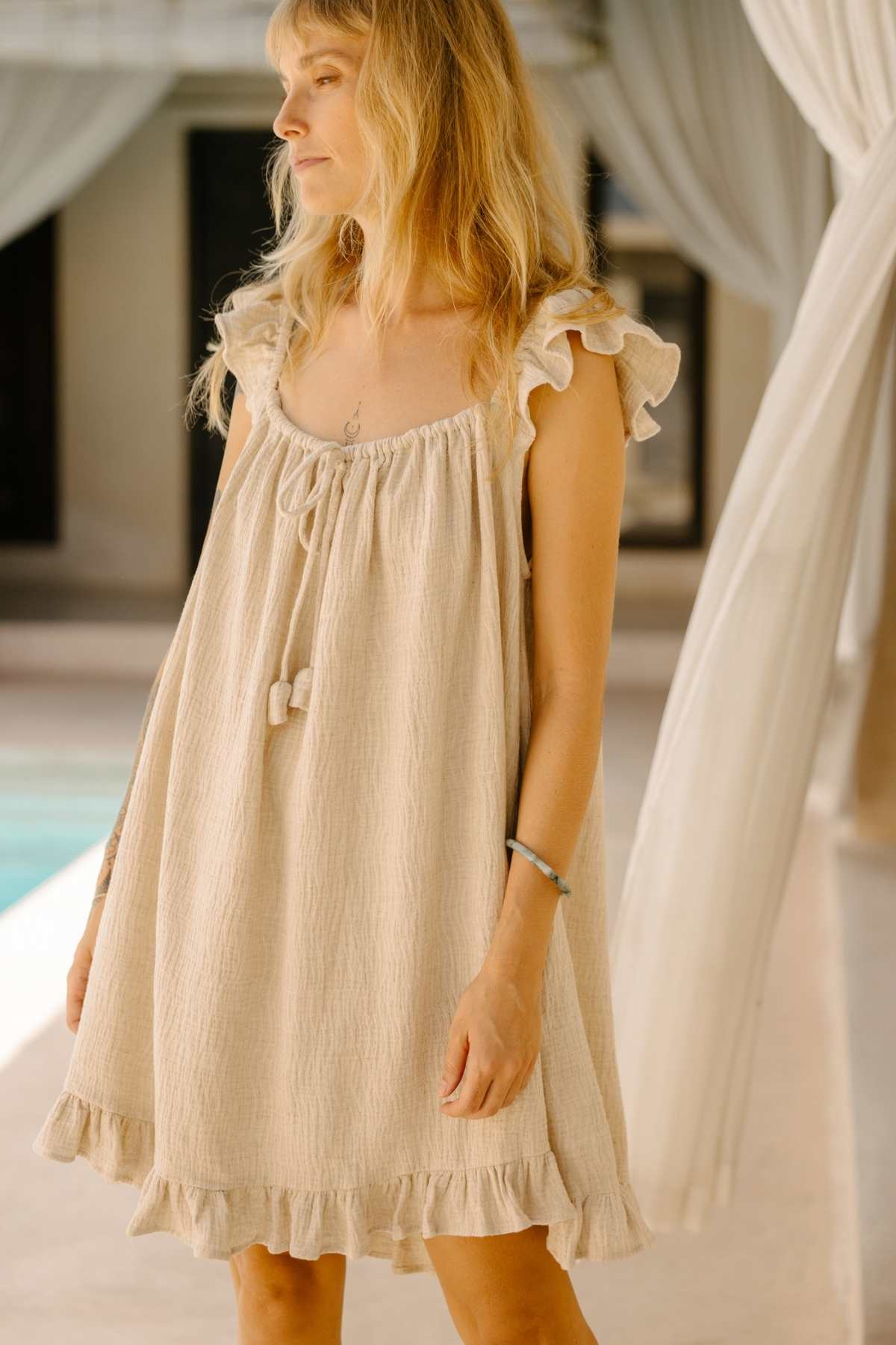 A woman with long blonde hair, wearing the light, sleeveless Bumi Mini Slip Dress by Myrah Penaloza stands indoors near a curtain and beside a pool, gazing off to the side with a relaxed expression. The setting appears serene and airy.