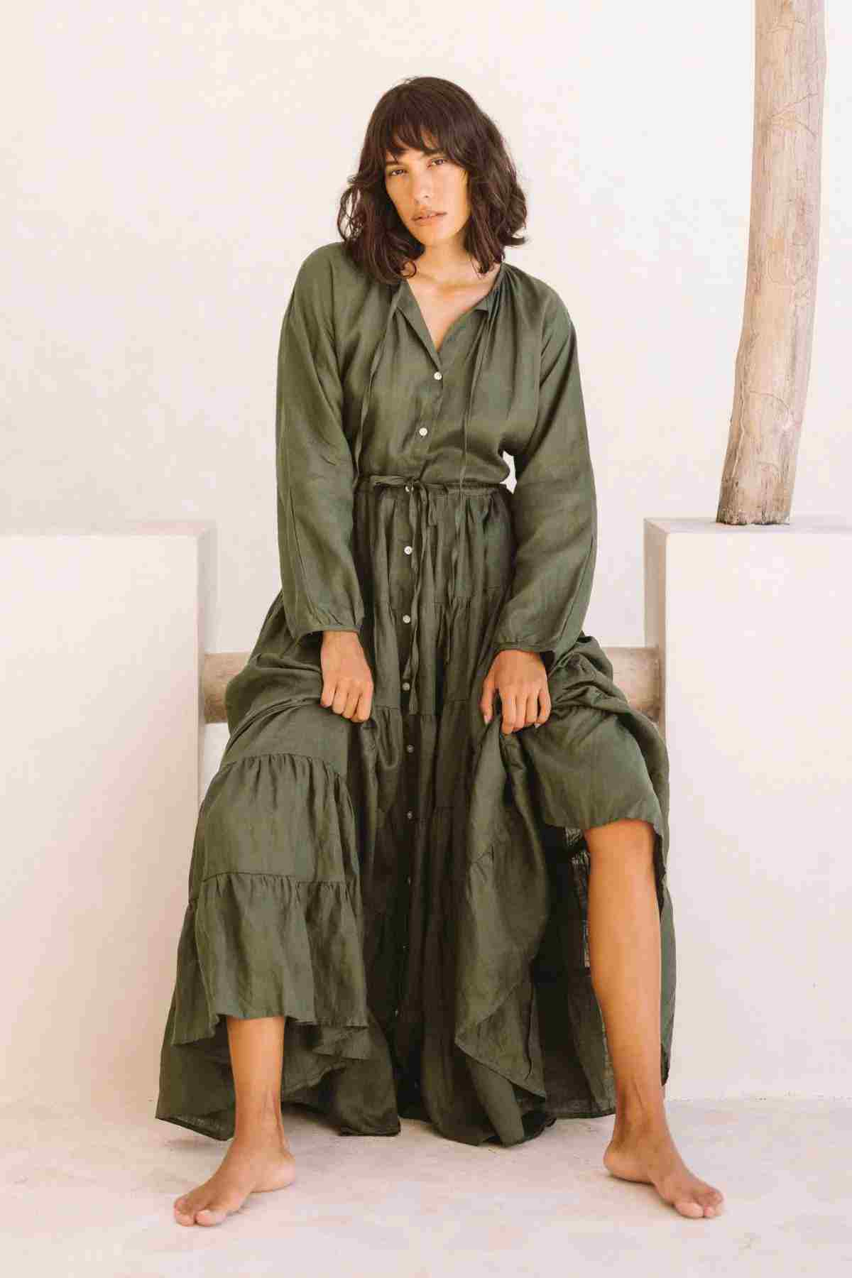 A woman sits gracefully on a bench, dressed in the La Mexicana Gown by Myrah Penaloza, featuring a long linen design in green with an adjustable waist. She is barefoot, one leg slightly raised, exuding effortless charm. The minimal backdrop showcases neutral tones and a wooden post framing the tranquil scene.