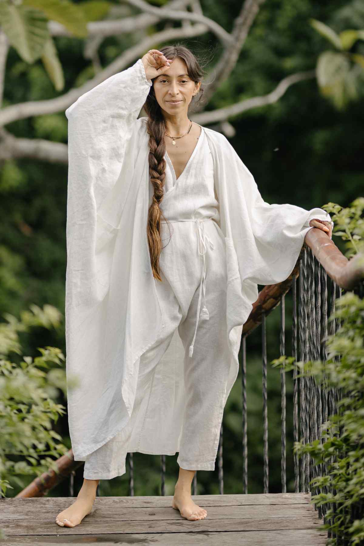 A barefoot person with long brown hair stands on a wooden bridge in a lush, green environment. Wearing the Myrah Penaloza Dao Robe (100% Linen) in natural colors, they hold the side of the bridge with one hand while shielding their eyes with the other.