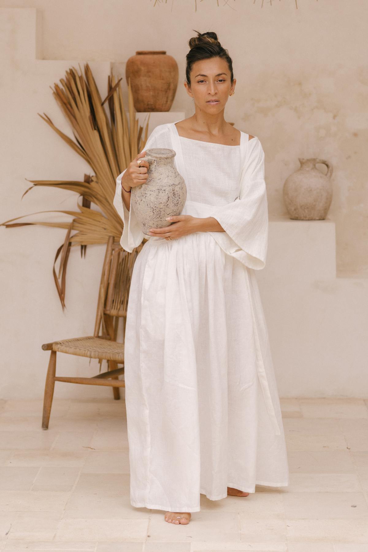 A woman dressed in a flowing, handmade May May Overall Skirt by Myrah Penaloza stands barefoot indoors, holding a large, weathered ceramic vase. She is positioned near a rustic wooden chair and dried palm fronds, against a textured beige wall with another vase in the background.