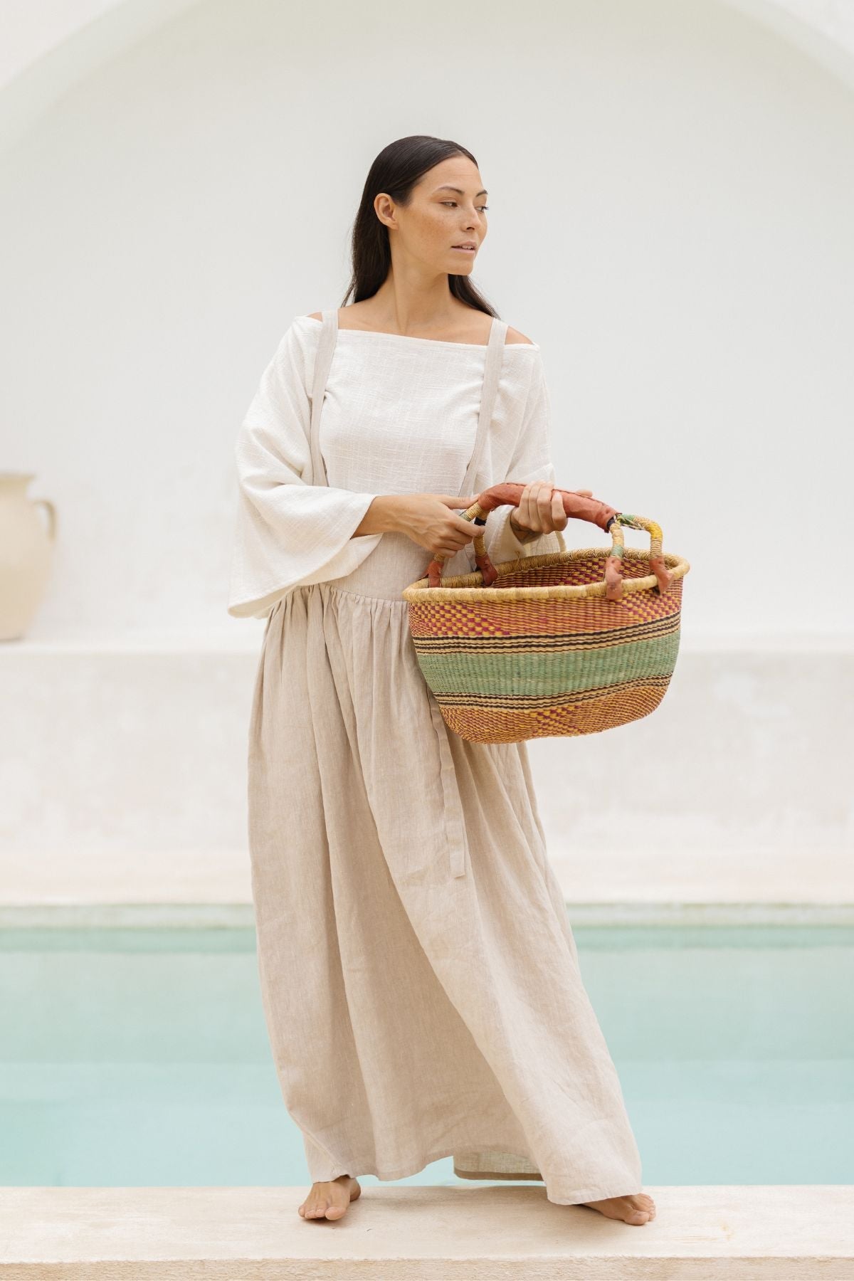 A woman with long dark hair is standing barefoot near a shallow pool. She is wearing the May May Overall Skirt (Made To Order) by Myrah Penaloza, which is crafted from 100% linen and has a light-colored design. She holds a large, multicolored woven basket in her hands. The background features a minimalist white arched structure.