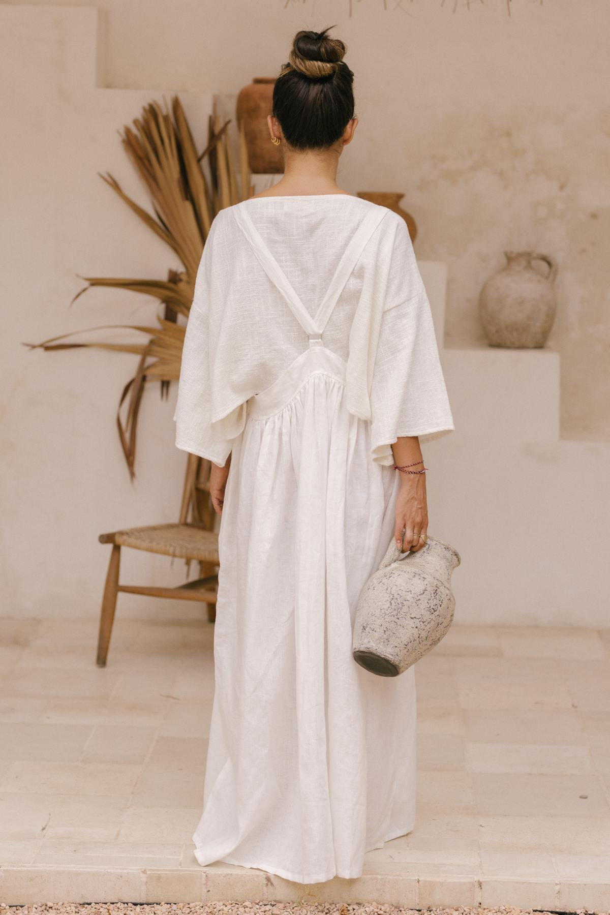 A person with their back to the camera stands in a neutral-toned room holding a large pottery vase. They are wearing the May May Overall Skirt (Made To Order) from Myrah Penaloza, which features a long, flowing white design made of crinkle linen with wide sleeves. Their hair is styled in a high bun. A wooden chair and dry plants are visible in the background.