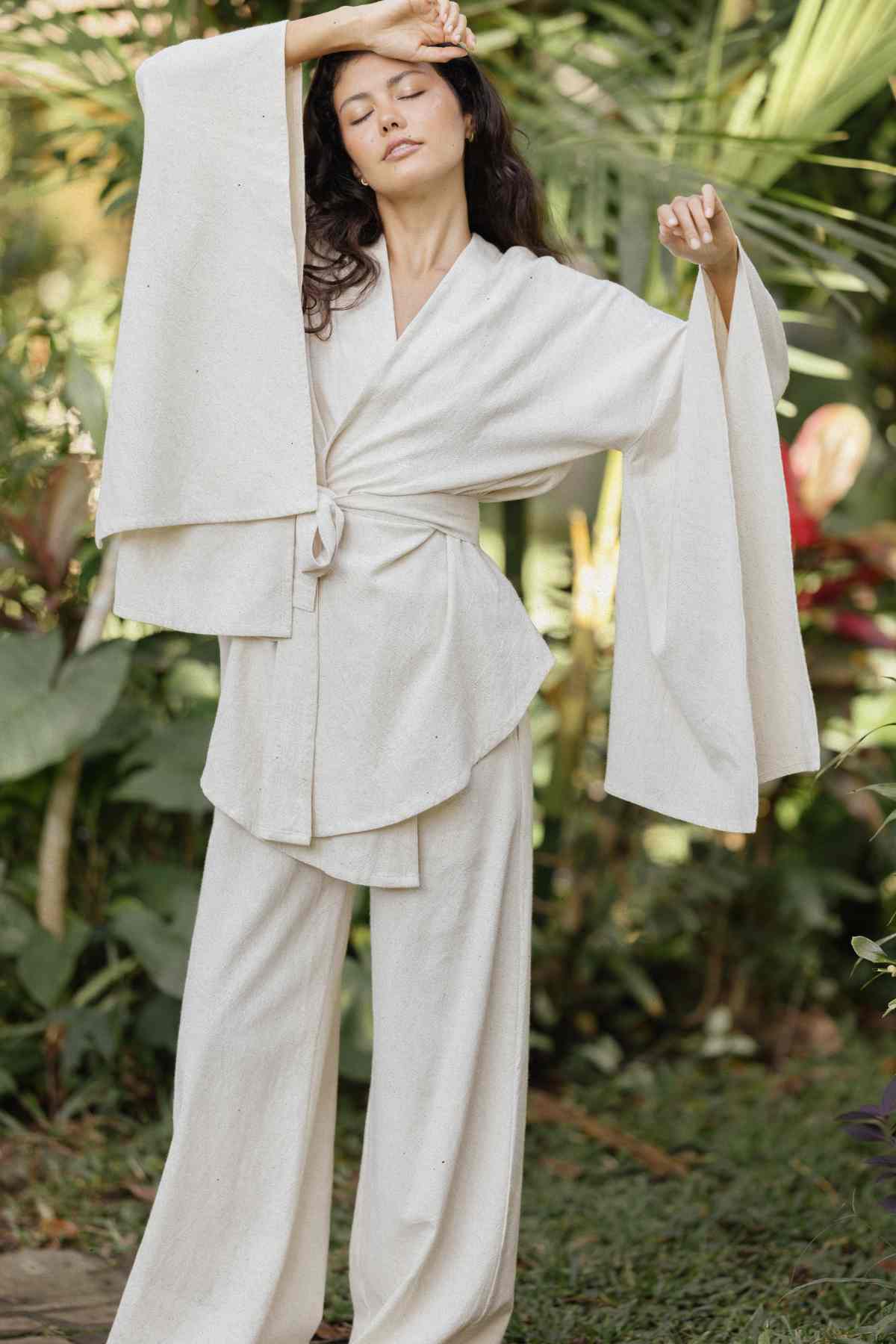 A person wearing the Dewi Set (3 of a kind) – Pre-order by Myrah Penaloza, a loose, light-colored kimono-style outfit crafted from hypoallergenic linen cotton crepe, poses outdoors with one arm raised and the other extended. They are surrounded by lush green foliage, conveying a sense of serenity and connection with nature.