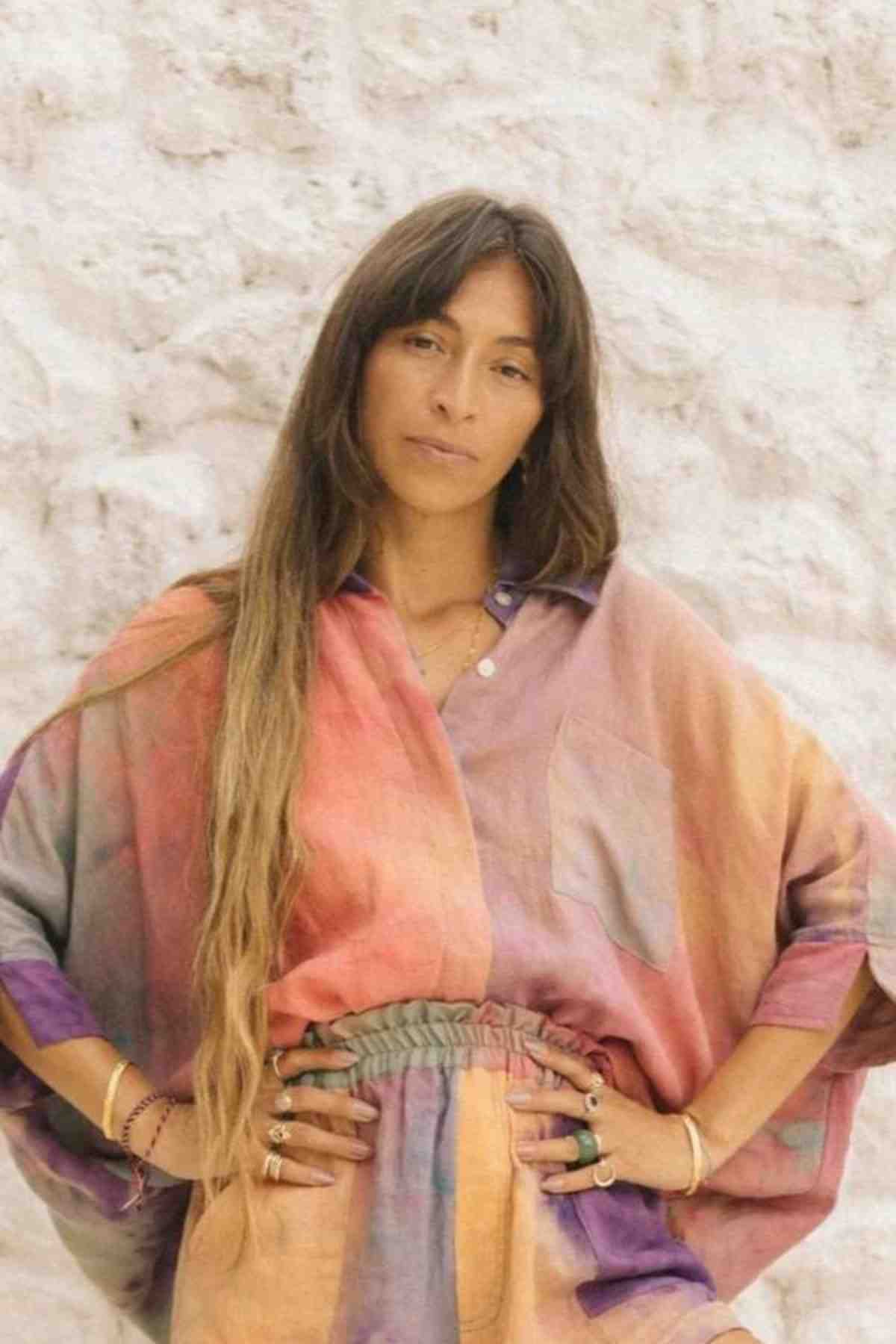 A person stands against a textured white wall, wearing the Suka Button Down Top from the Rainbeau Collection by Myrah Penaloza, made of 100% sustainable linen. The long sleeves and tastefully added rings complement their flowing hair. With hands on hips, they exude a calm confidence that reflects the allure of ethically sourced fashion.