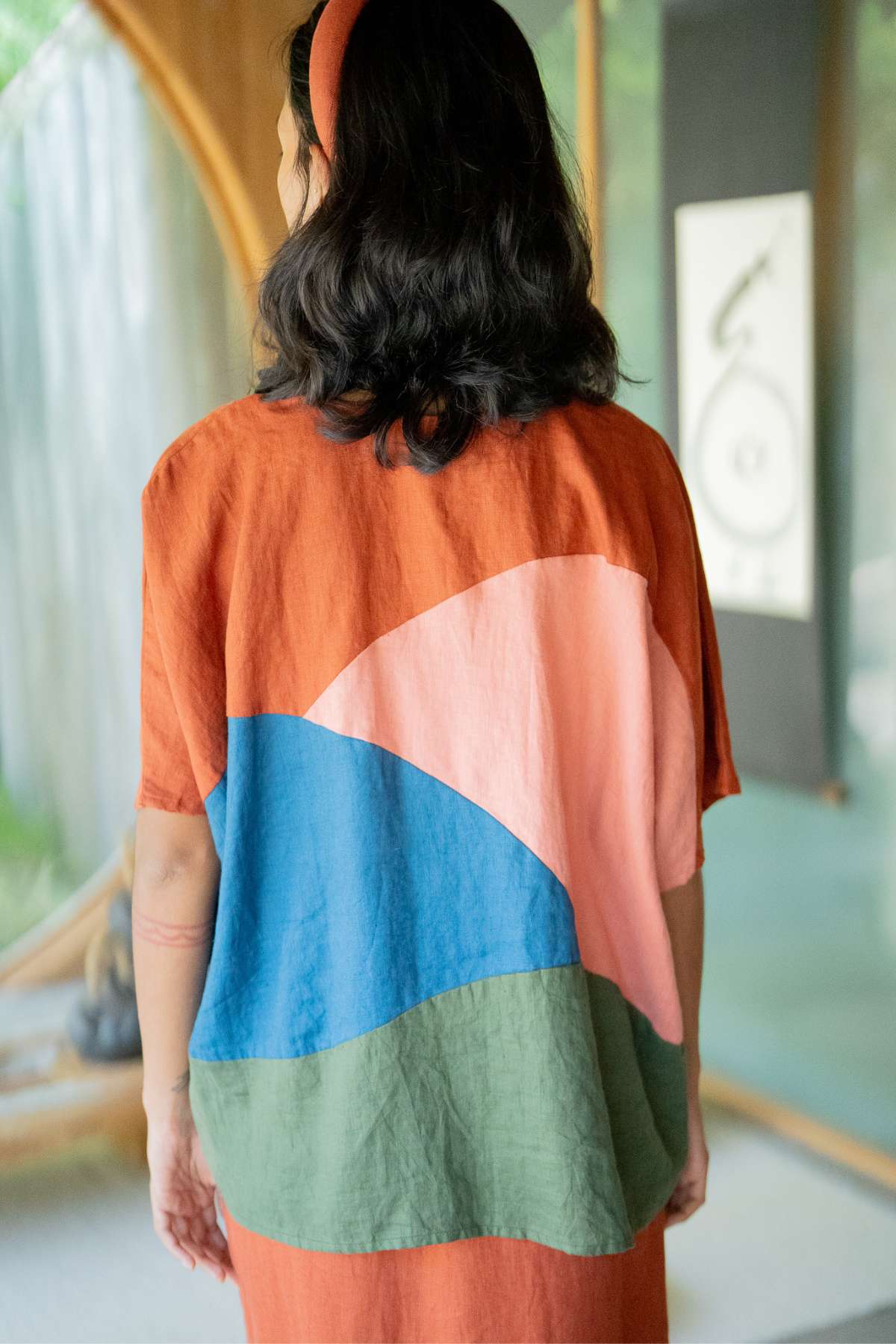 A person with wavy hair, wearing an Uluwatu Top by Myrah Penaloza and an orange dress adorned with colorful patterns in pink, blue, and green, stands indoors near a large circular window.