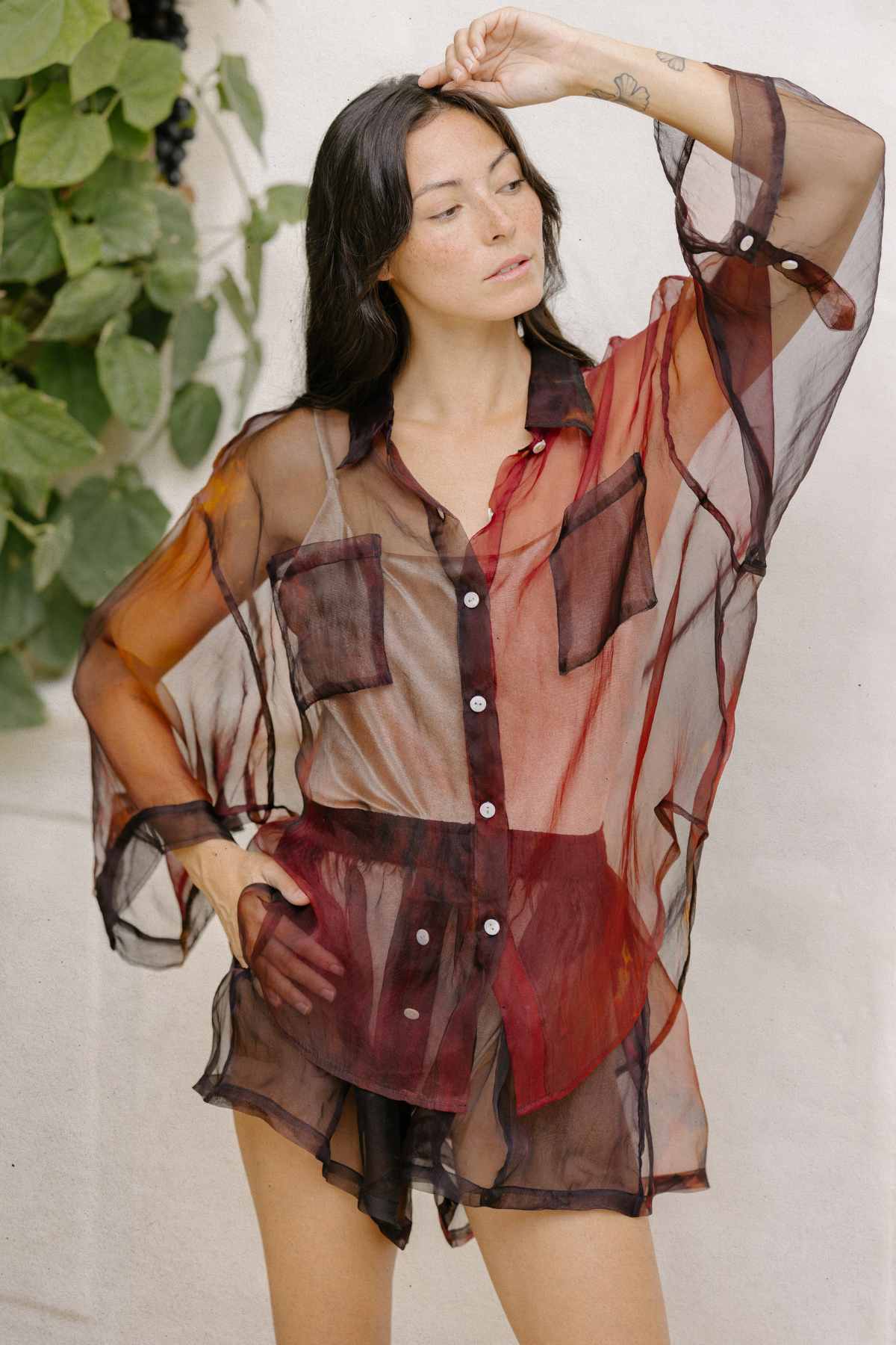 A person with long dark hair is posing outdoors wearing the Rainbeau Nidra Chiffon Set from Myrah Penaloza. The sheer, flowing blouse has long sleeves, a pocket, and button-down features. They have one arm raised and resting on their head, with green foliage visible in the background.