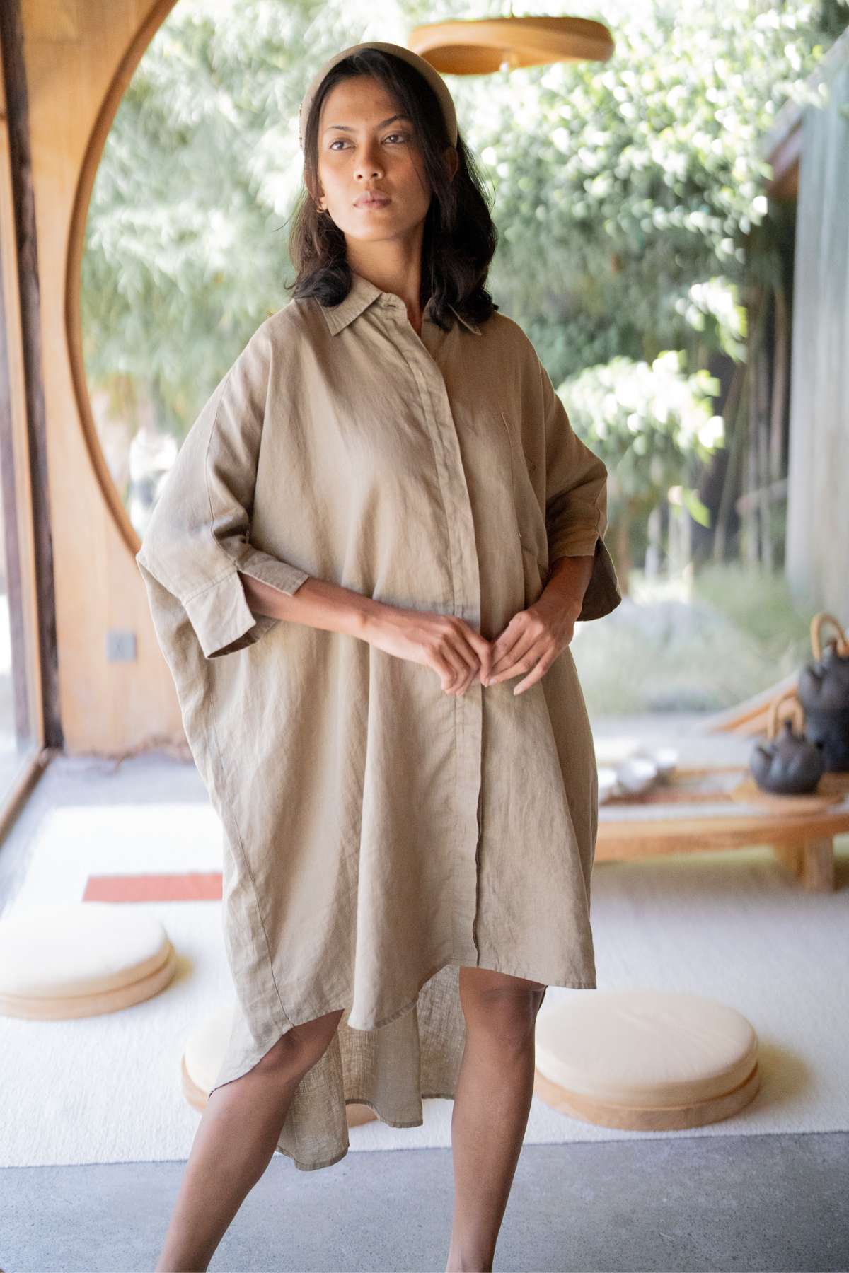 An individual stands indoors wearing the Linen Suka Button Down Long Top - BF by Myrah Penaloza, made from eco-friendly materials. They are positioned before a large circular window overlooking lush greenery. The soft lighting and minimalist decor create a tranquil atmosphere, showcasing the top's versatile styling options.