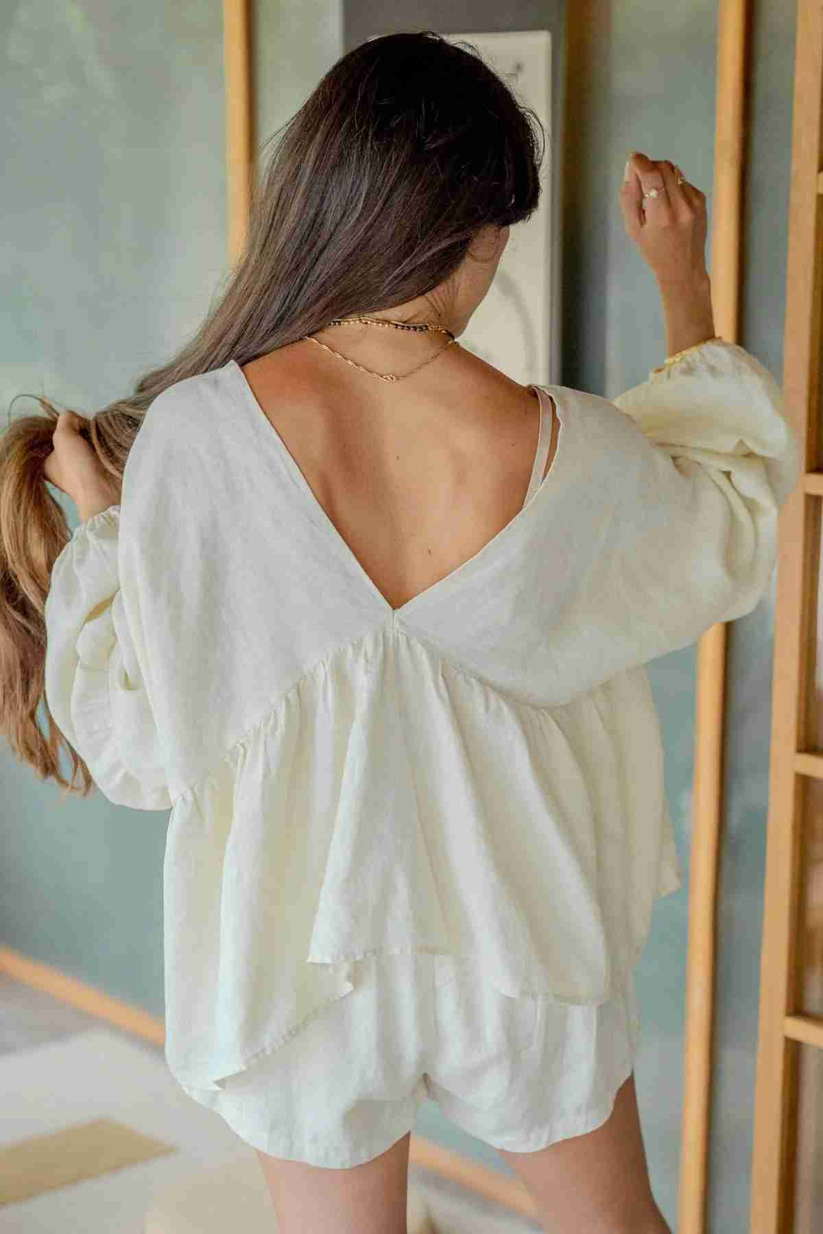 A woman with long brown hair, seen from behind, is wearing a loose-fitting Eva Linen Top by Myrah Penaloza with a deep V-back and a matching white skirt. She is standing in an interior space adorned with light blue and wooden accents while casually holding her hair with one hand.