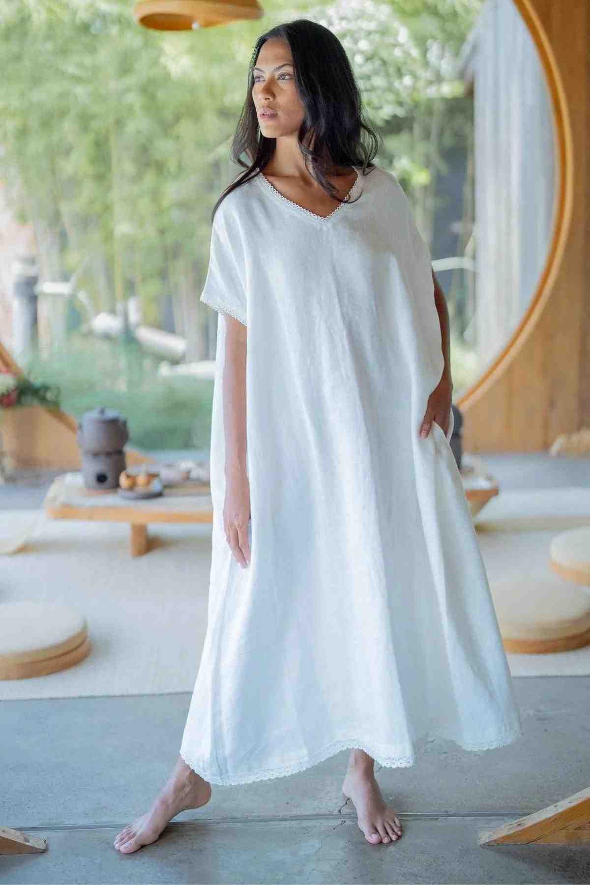 A woman in a flowing, V-neckline Ruby Lace Tunic from Myrah Penaloza stands barefoot on a concrete floor inside a serene room with large circular windows. She gazes to the side with a relaxed expression while bamboo plants and a low wooden table with teaware provide a tranquil backdrop.