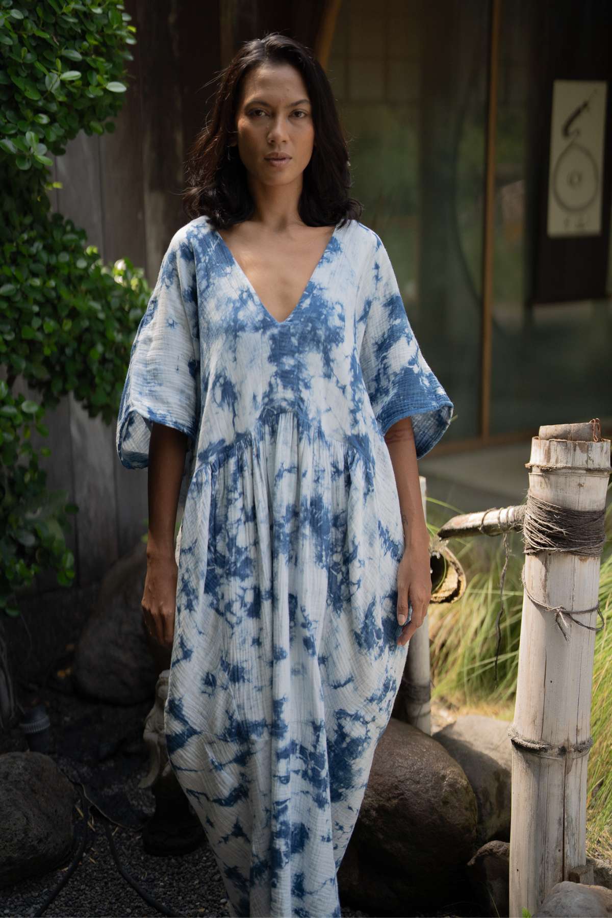 A person stands outdoors enveloped in a flowing, effortless style, wearing the Indigo Venus Gown by Myrah Penaloza. Surrounded by greenery and a wooden fence, they embody Venus in her natural element, with part of a building featuring a wall painting in the background.