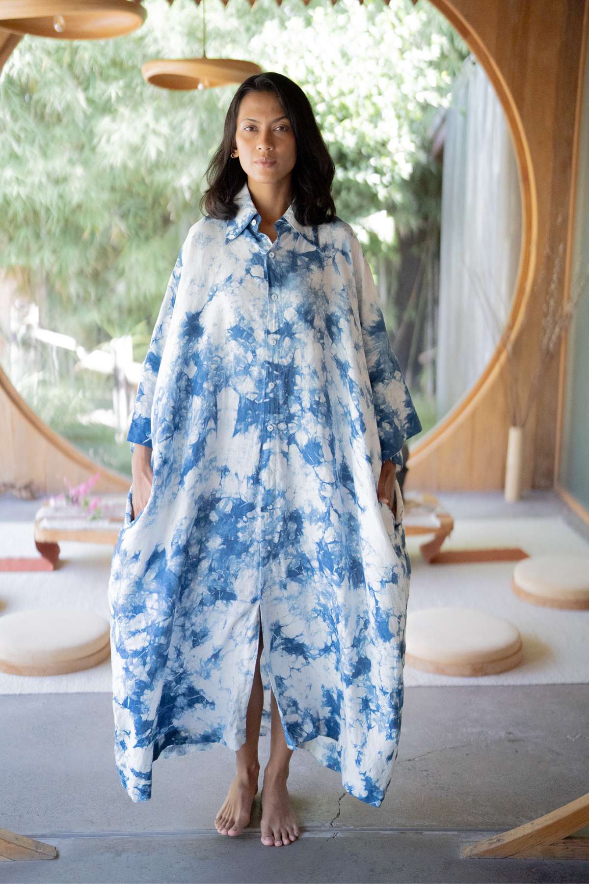 A woman stands barefoot in a room, wearing The Boyfriend Dress by Myrah Penaloza, hand tie-dyed in indigo and reminiscent of a Kundalini gown. The space features a large round window with a lush garden view. She holds the dress at the sides, as natural light illuminates the serene setting, enhancing its tranquil ambiance.