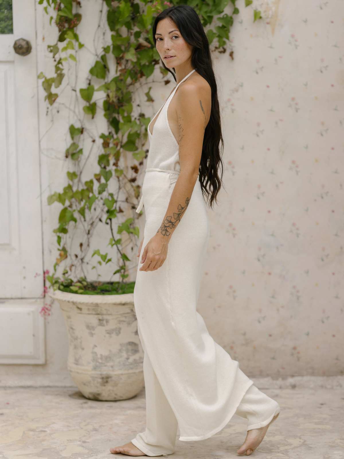 A woman with long dark hair is wearing a Honey Cotton Knit Wrap Dress by Myrah Penaloza. She stands sideways, looking at the camera, in front of a white door with vines climbing up the wall. The floor is made of stone, and there's a large potted plant beside her.