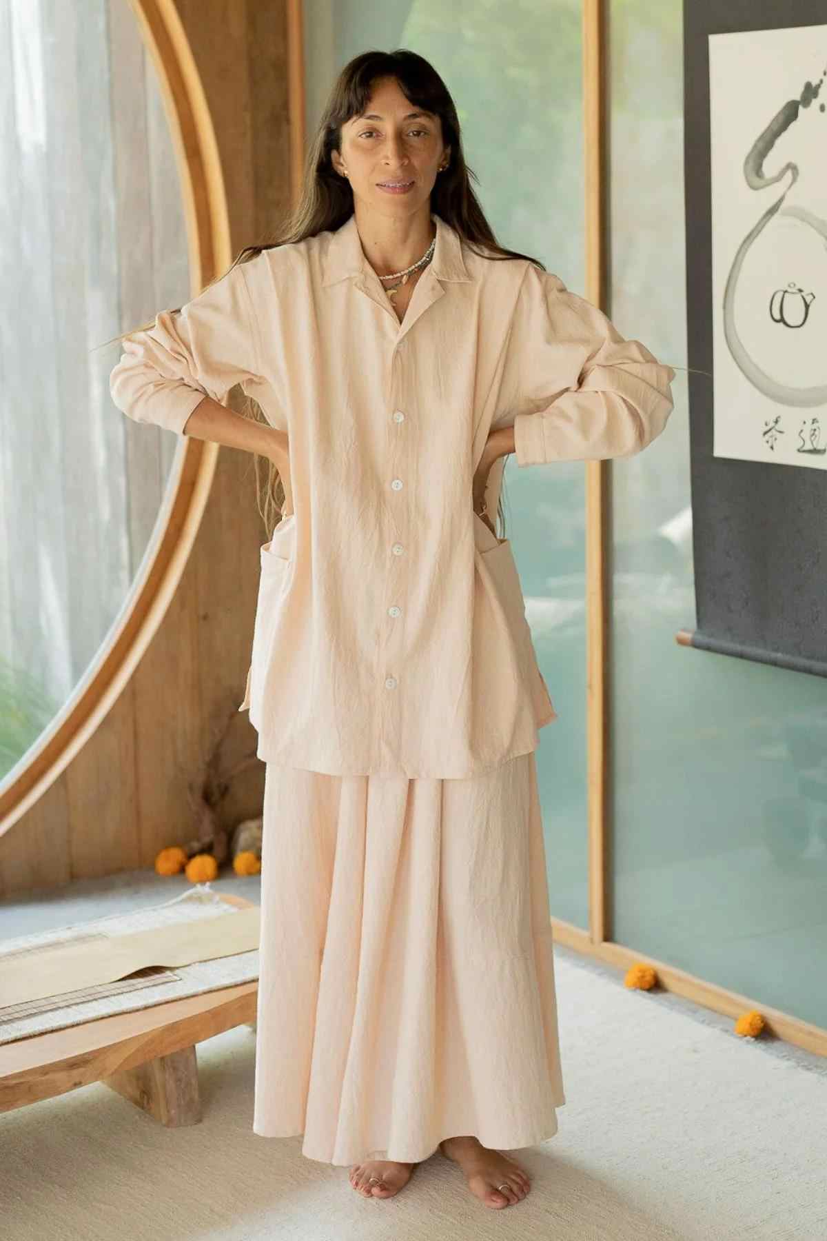 A woman stands indoors with her hands on her hips, wearing the Tender Jacket & Pant Set (3 of a kind) from Myrah Penaloza. She has long dark hair and is barefoot. The background shows a large circular window and a piece of wall art.