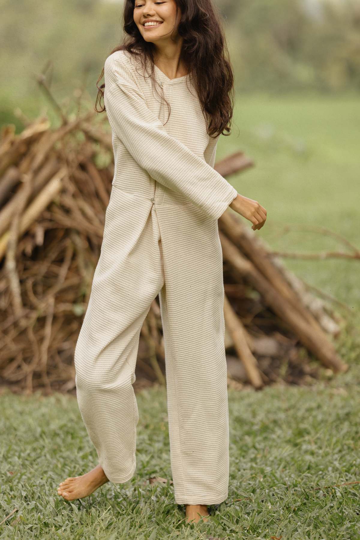 A person with long dark hair, dressed in a loose-fitting Myrah Penaloza Camper Playsuit made from waffle cotton, is standing outside on the grass, smiling and moving slightly. In the background, there's a pile of sticks and greenery, creating a relaxed and natural atmosphere.