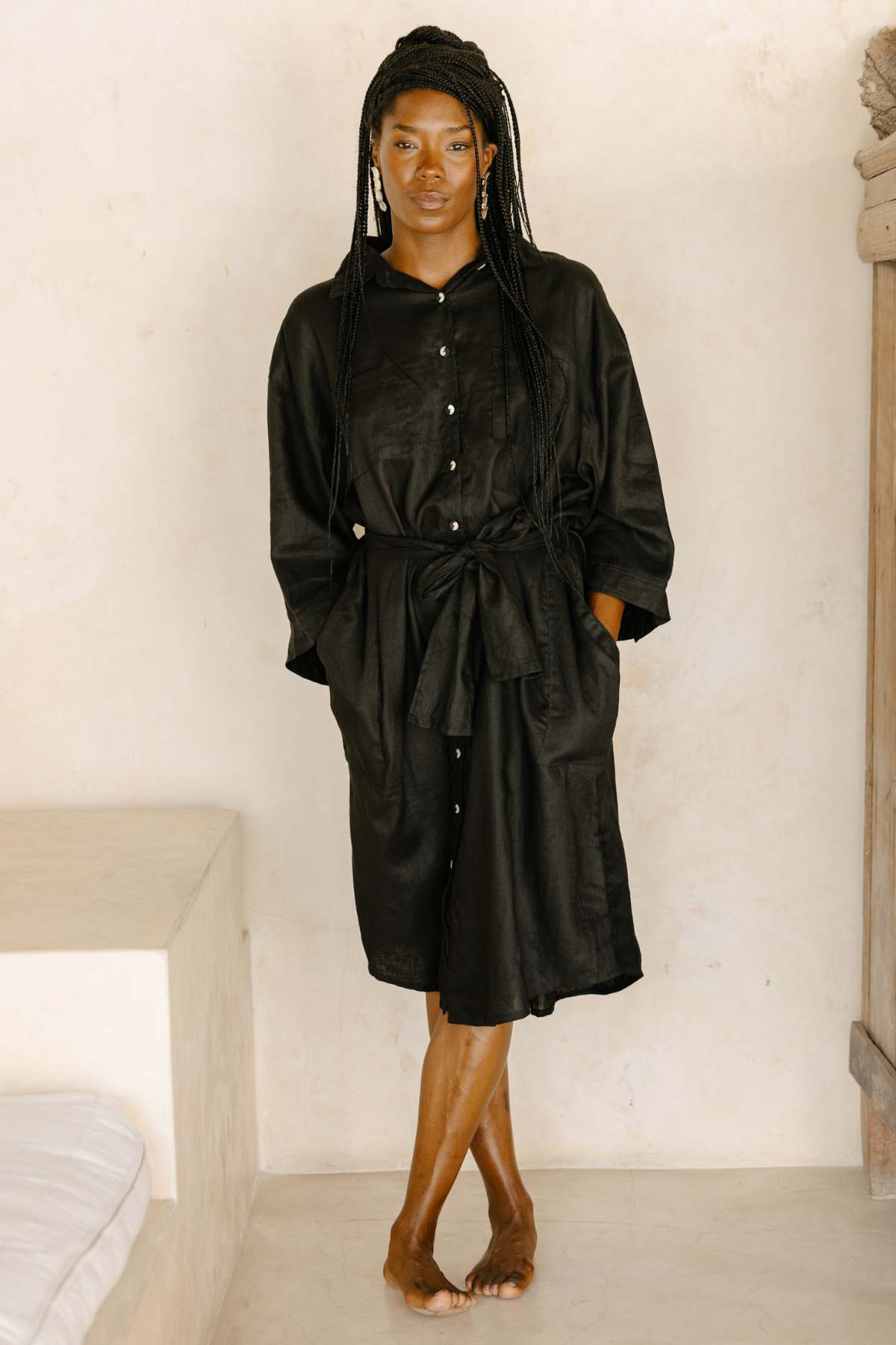 A person gracefully leans barefoot against a beige wall indoors, wearing the Nidra Full Length Linen Duster by Myrah Penaloza. The black duster features a waist tie and exudes sustainability, perfectly complementing their long braided hair and serene expression. In this minimalist space accented with neutral tones, the oversized fit and eco-friendly fabric choice of the duster make a subtle yet profound statement.