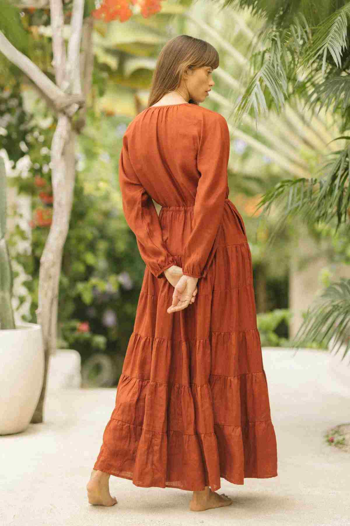 A person stands barefoot on a stone path, wearing the long La Mexicana Gown in rust color by Myrah Penaloza. They face away with hands clasped behind their back, enveloped by lush greenery and plants.