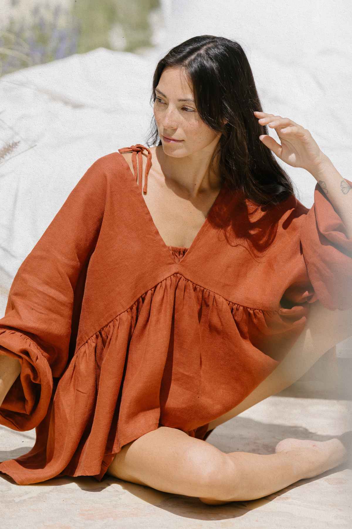 A woman with long dark hair is sitting outdoors, wearing an orange Eva Linen Top by Myrah Penaloza, with loose, flowing sleeves. She has a relaxed pose, with one hand tucking her hair behind her ear. The background is softly blurred with hints of greenery and light fabric.