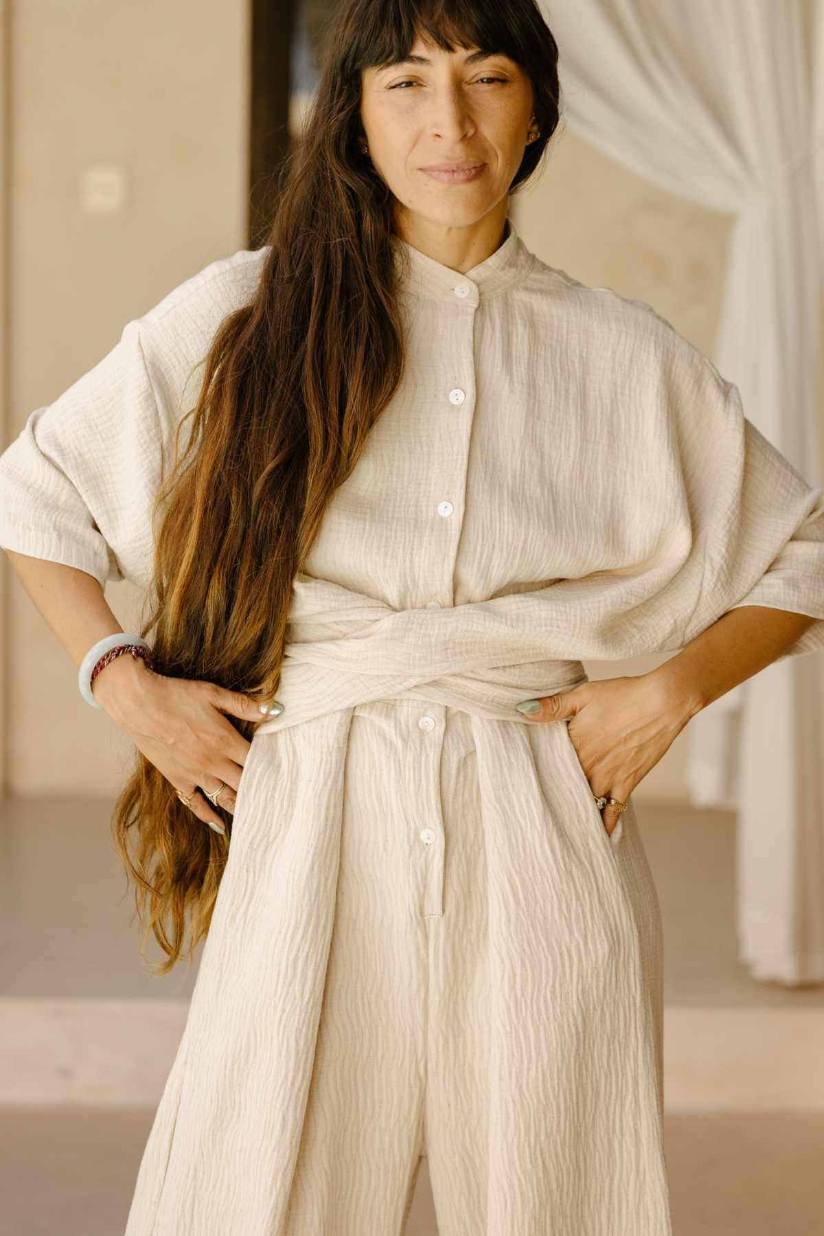 A person with long hair stands confidently in the Pine Playsuit by Myrah Penaloza, hands on hips, in a softly lit room with light curtains in the background. This neutral-toned romper, crafted from a cotton/linen blend, exudes effortless style and comfort as a versatile wardrobe piece.