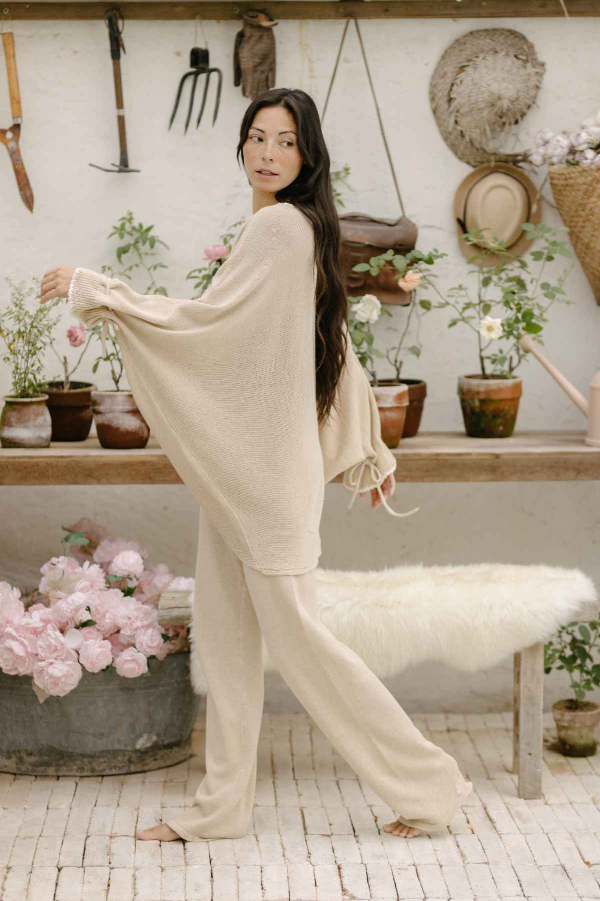 A woman with long dark hair, dressed in a Honey Knit Sweater by Myrah Penaloza and matching loose pants, stands barefoot in a rustic room filled with potted plants and gardening tools. She gazes to the side, while behind her, a wooden table adorned with plants and a straw hat hanging on the wall completes the serene scene.